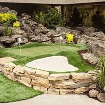 Small backyard putting green with a sand bunker, rock border, and two water sprinklers.