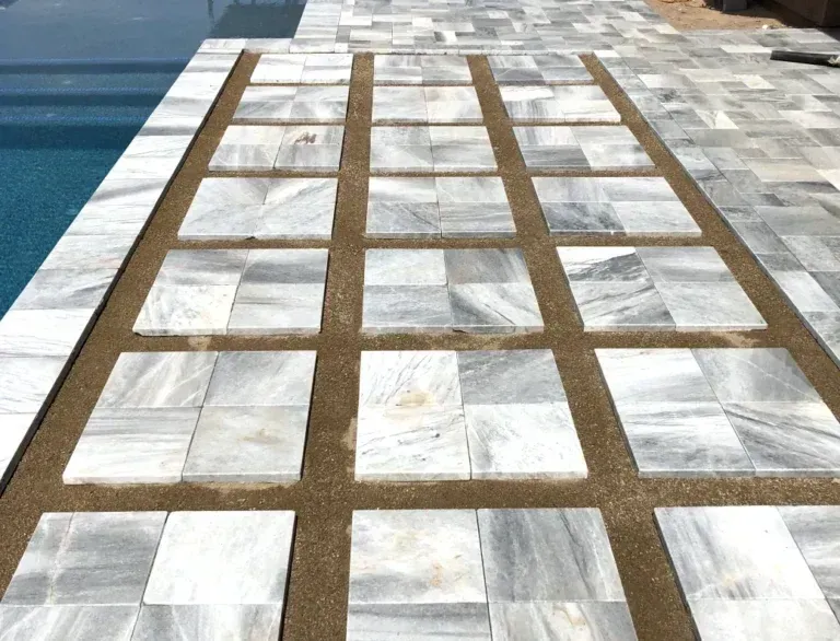White and gray tiled walkway with brown grid lines beside a blue pool or waterfront.
