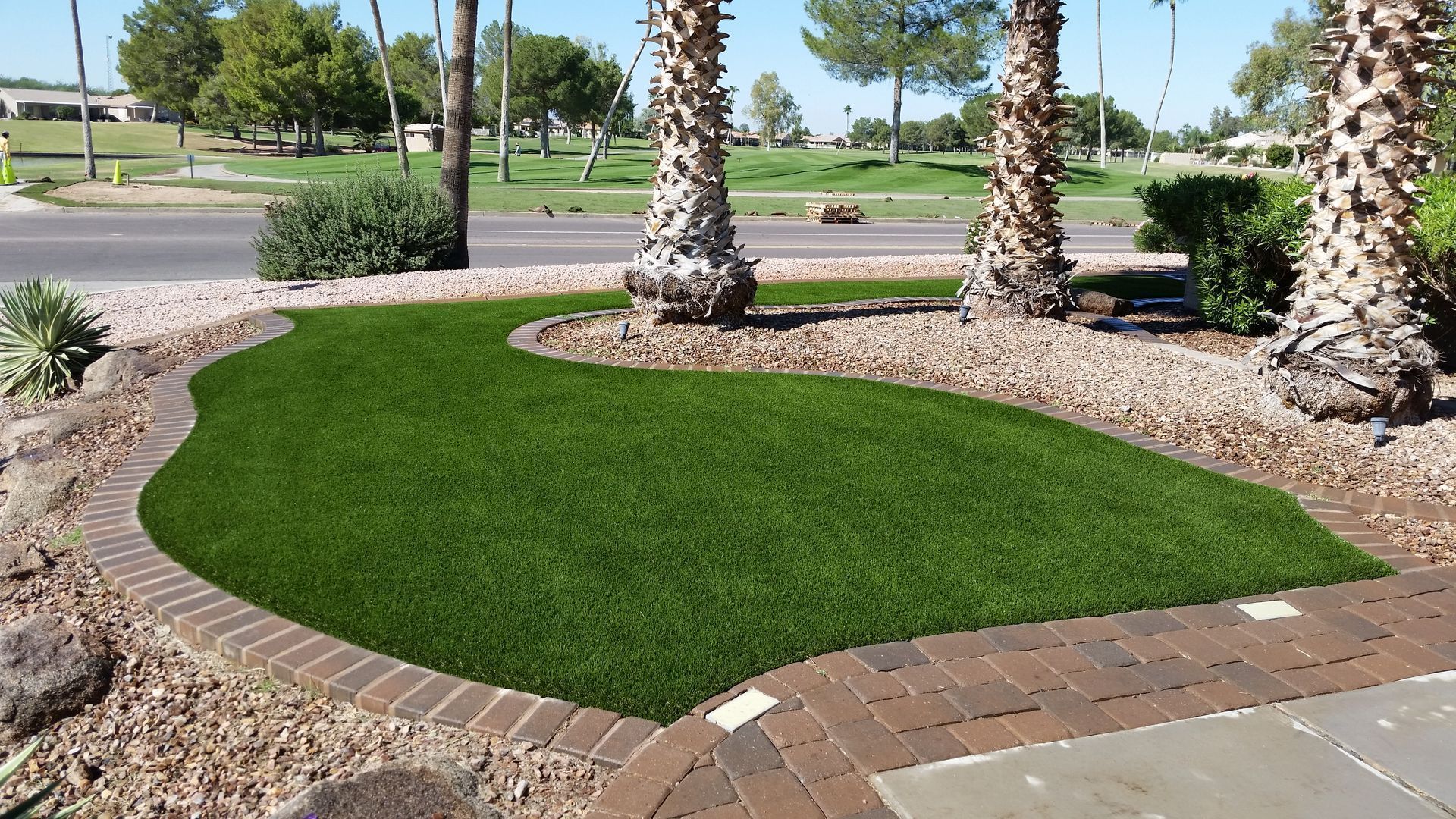 Curved patch of artificial green turf bordered by rocks and pavers between palm trees in a sunny landscaped area