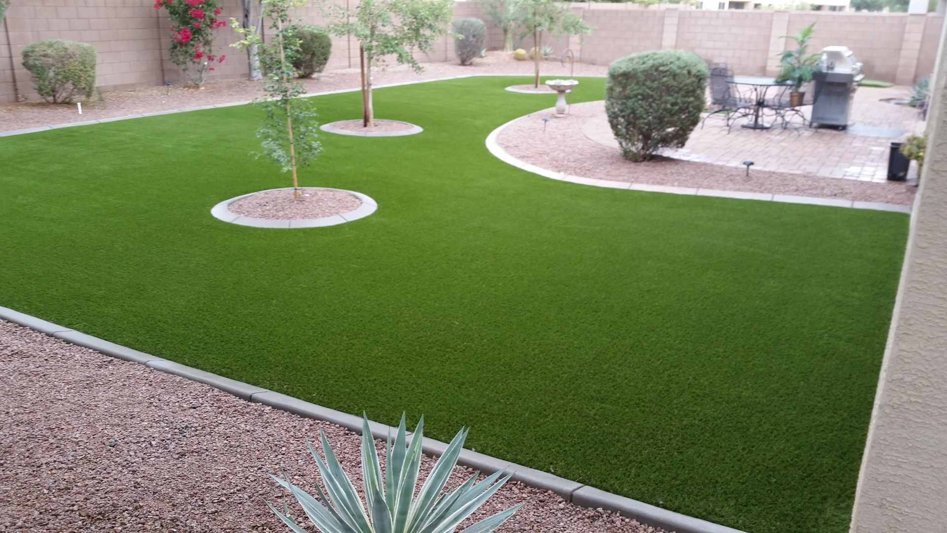 Landscaped backyard with bright green lawn, young trees, gravel borders, and a patio seating area