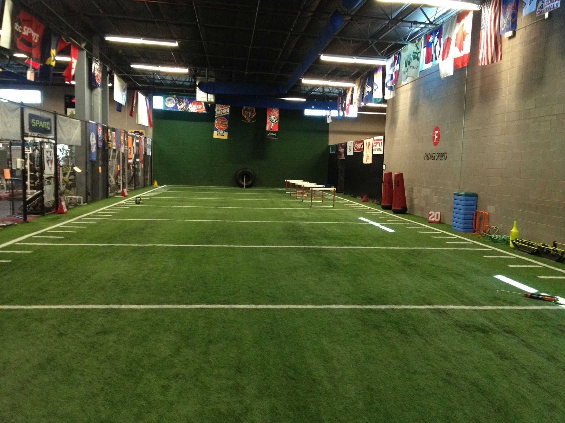 Indoor turf field with white yard lines and flags, viewed from one end toward a green back wall.
