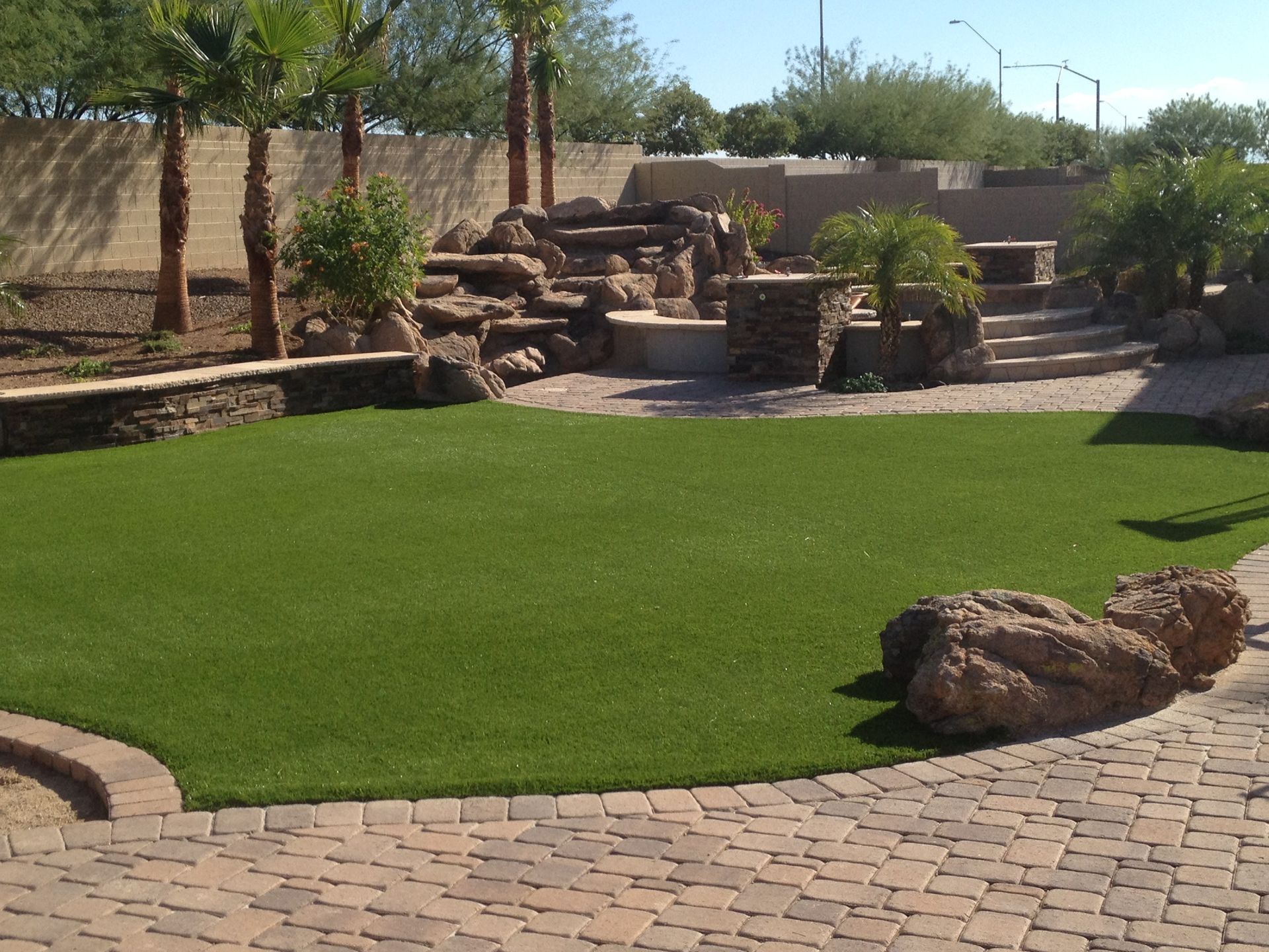 Desert garden with green lawn, stone fountain, palm trees, and brick walkway under a clear sky