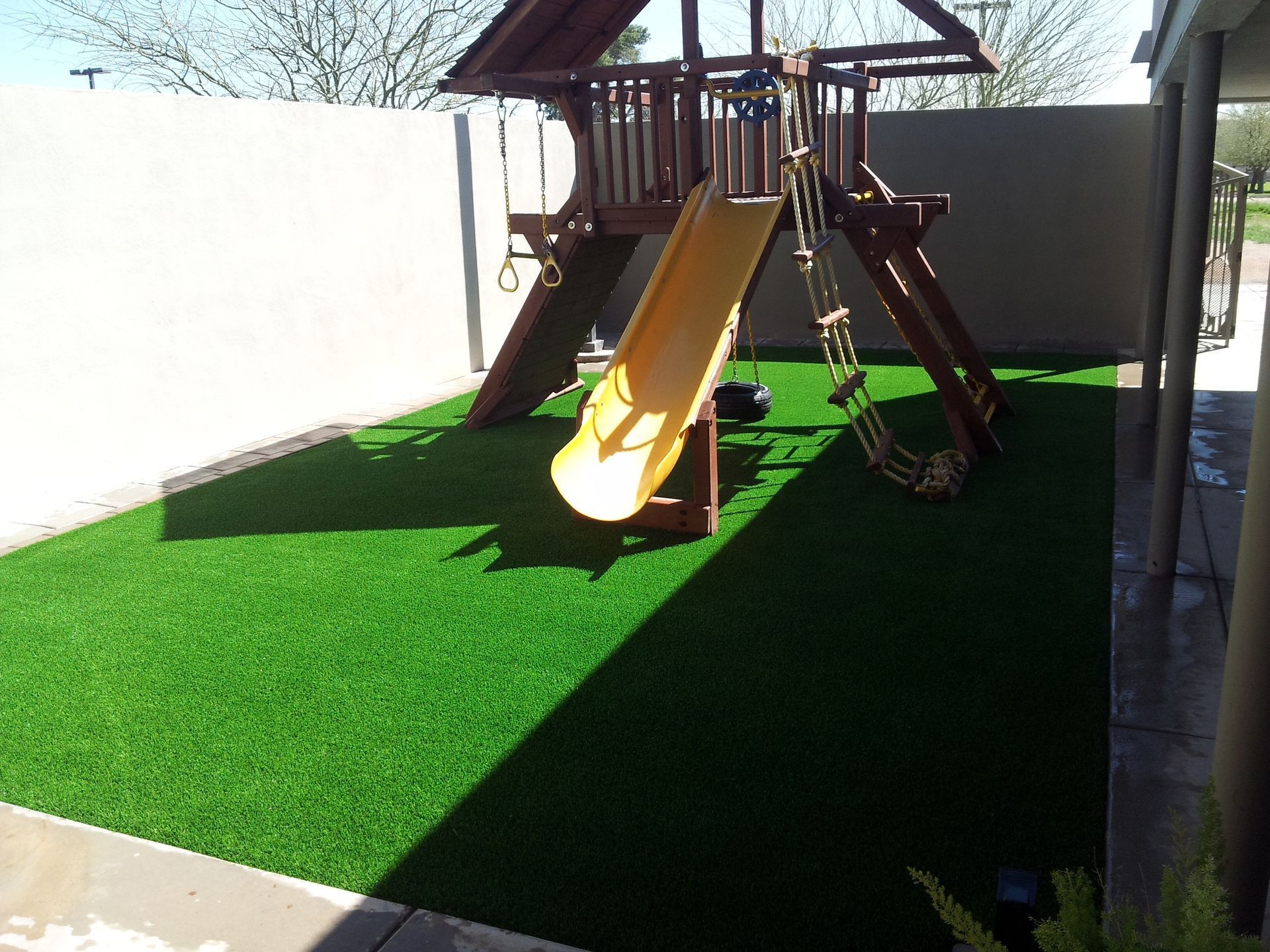 Backyard playground with a yellow slide on bright green artificial turf, enclosed by white walls.