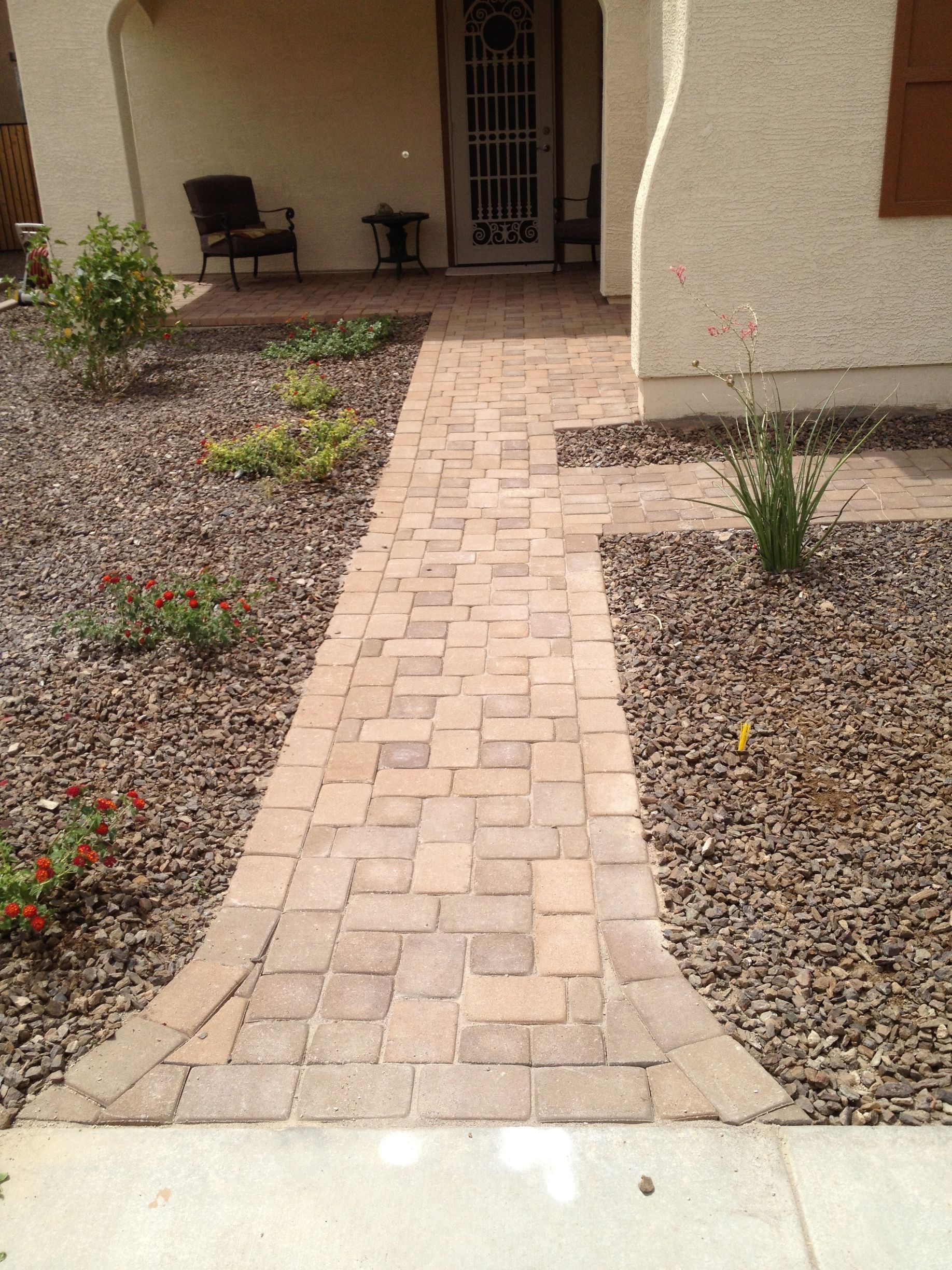 Brick walkway leading to a front porch with chairs and potted plants, bordered by gravel landscaping.