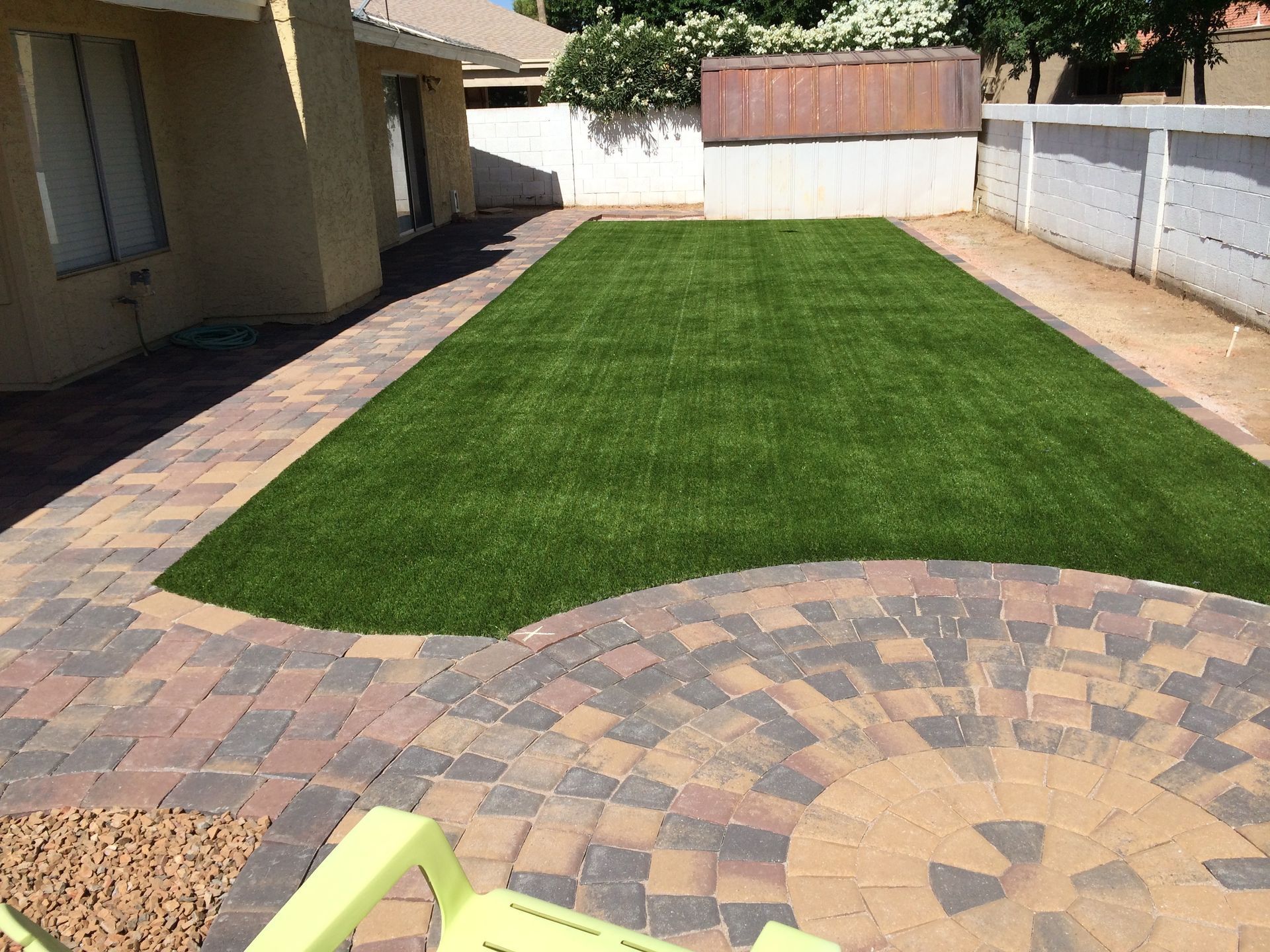 Backyard with a green lawn bordered by tan pavers and a white fence beside a house