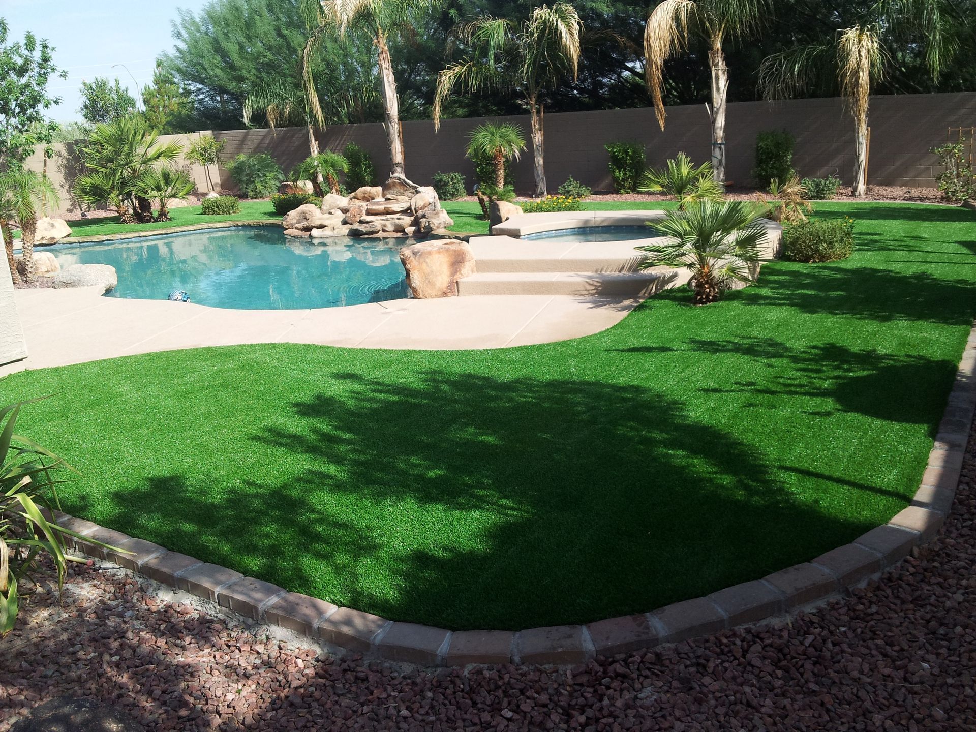 Backyard pool with rock waterfall, palm trees, and lush green lawn on a sunny day