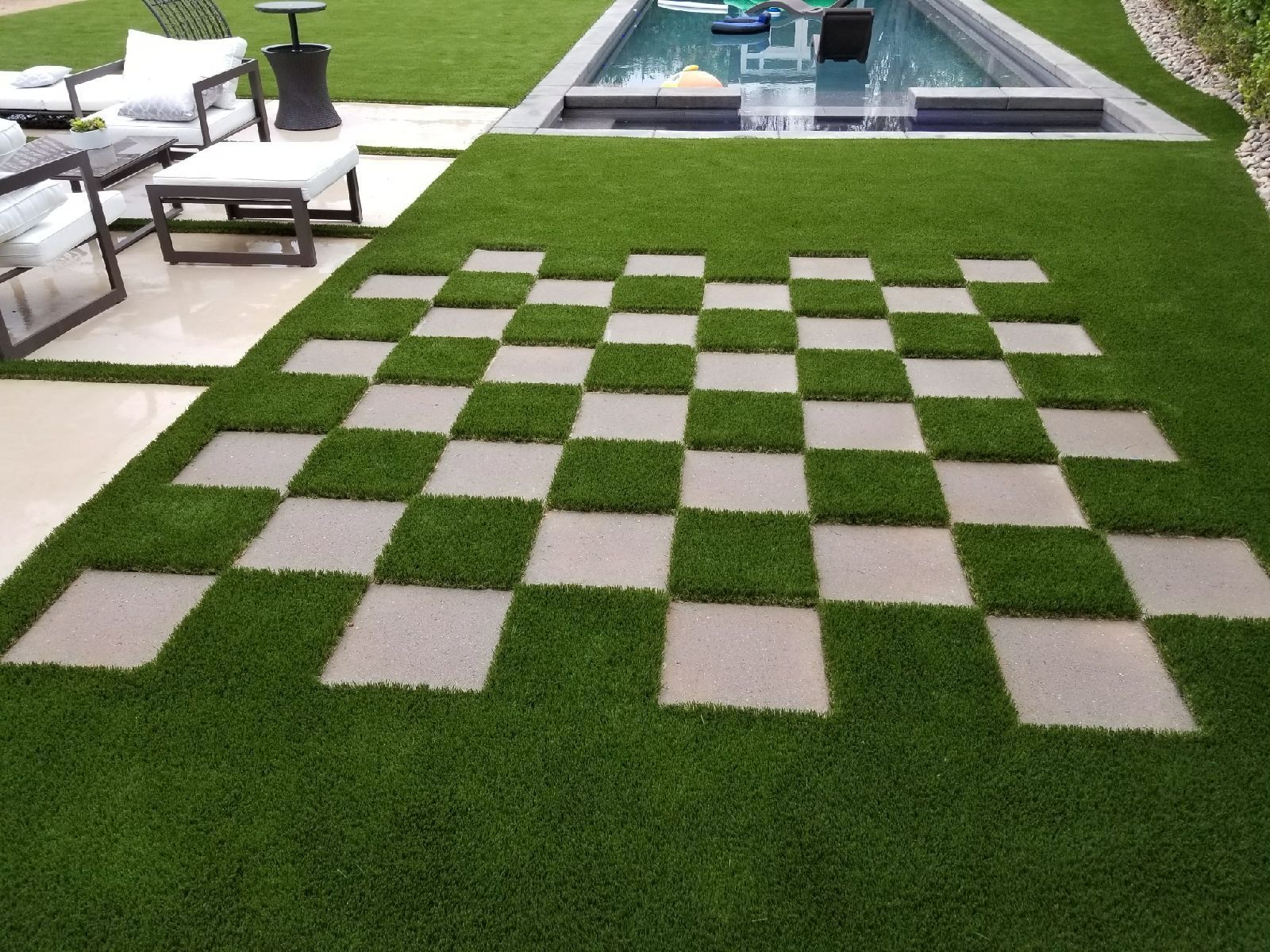 Checkerboard-style stone pavers set in green grass beside a modern pool and patio furniture.