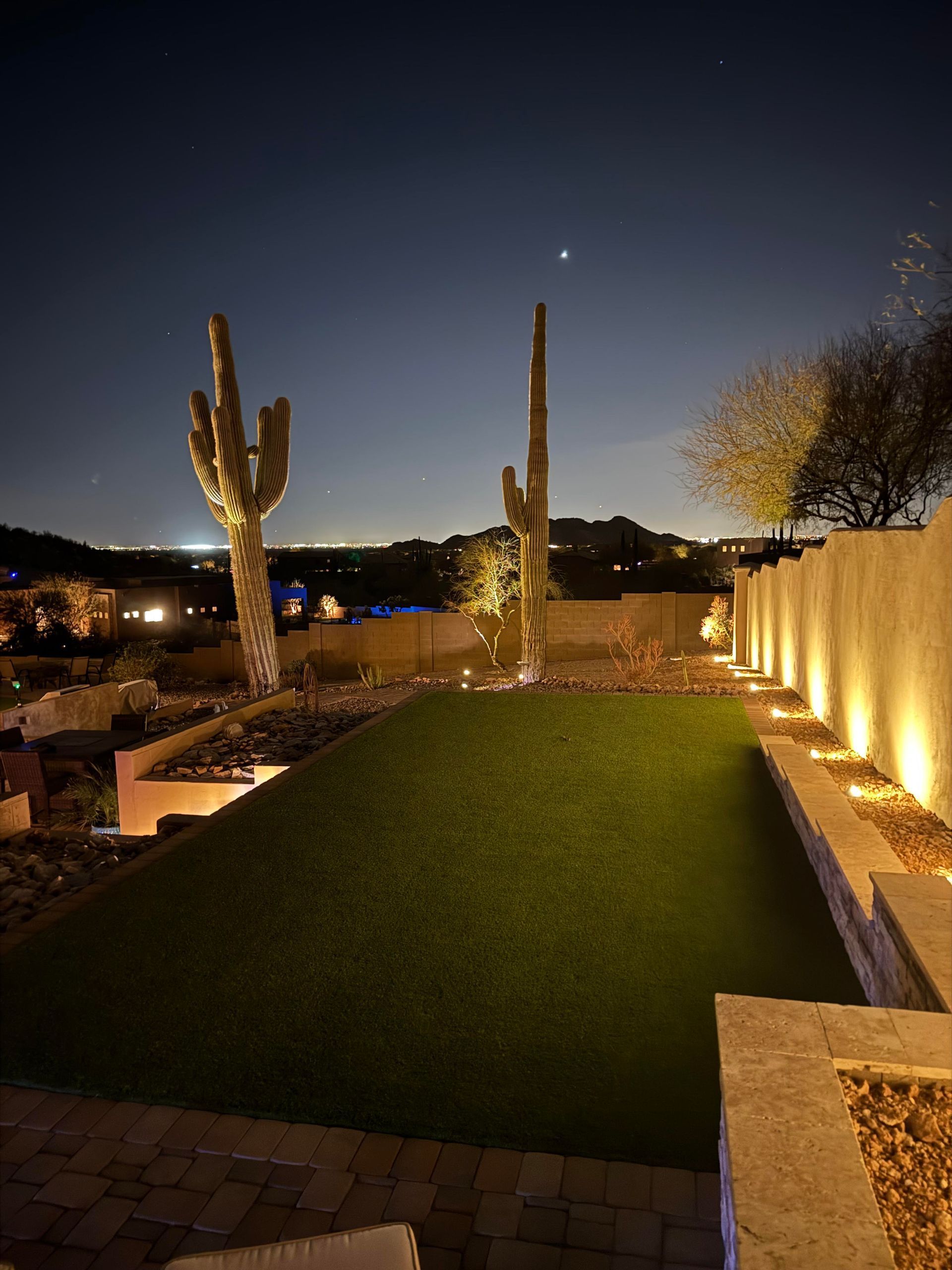 Nighttime desert courtyard with lit cacti sculptures, green lawn, and warm wall lights under a dark sky