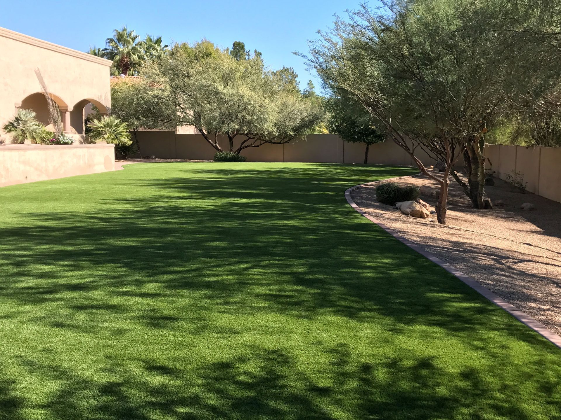 Sunny backyard lawn with green grass, trees, and a stucco house beside a gravel path