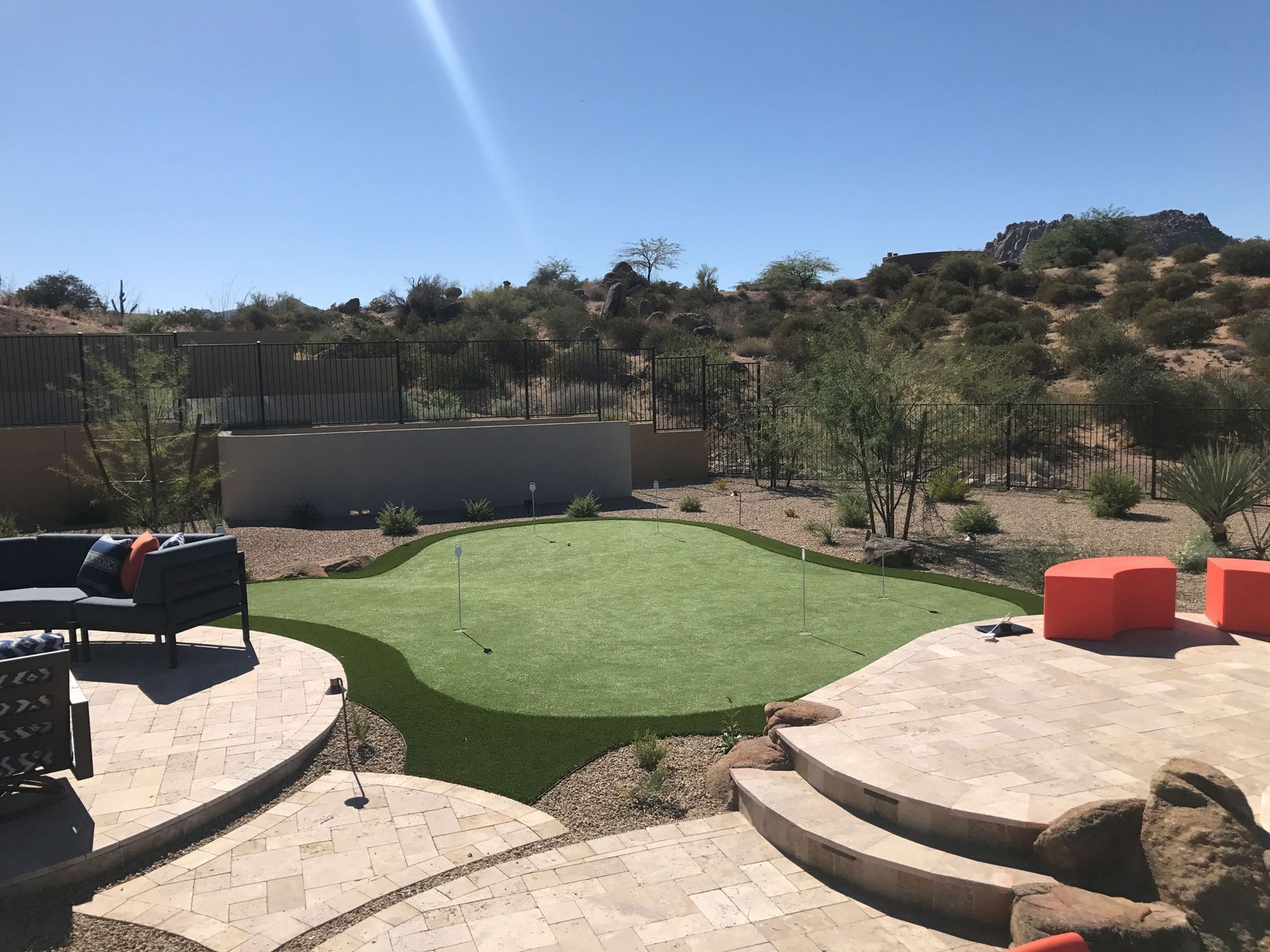 Backyard patio with curved stone steps, artificial turf, outdoor seating, and desert landscaping under a blue sky
