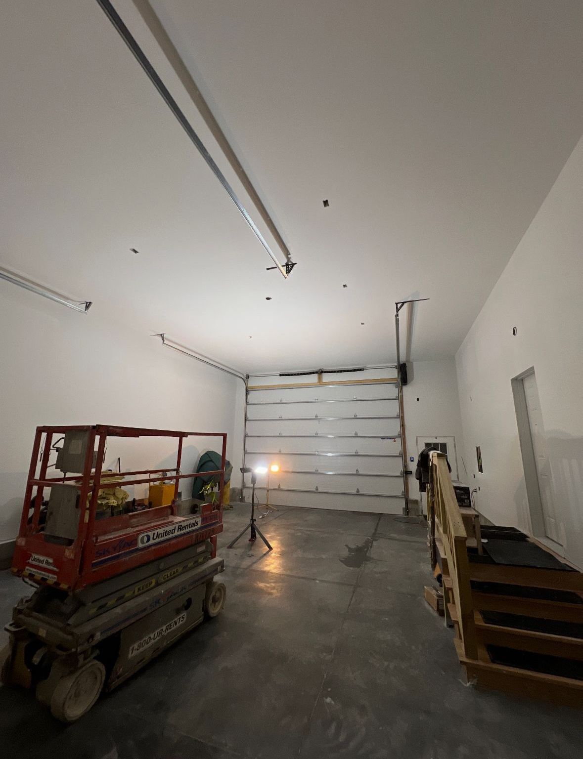 Interior of a white-walled warehouse with a red scissor lift, overhead garage door, and bright work lights.
