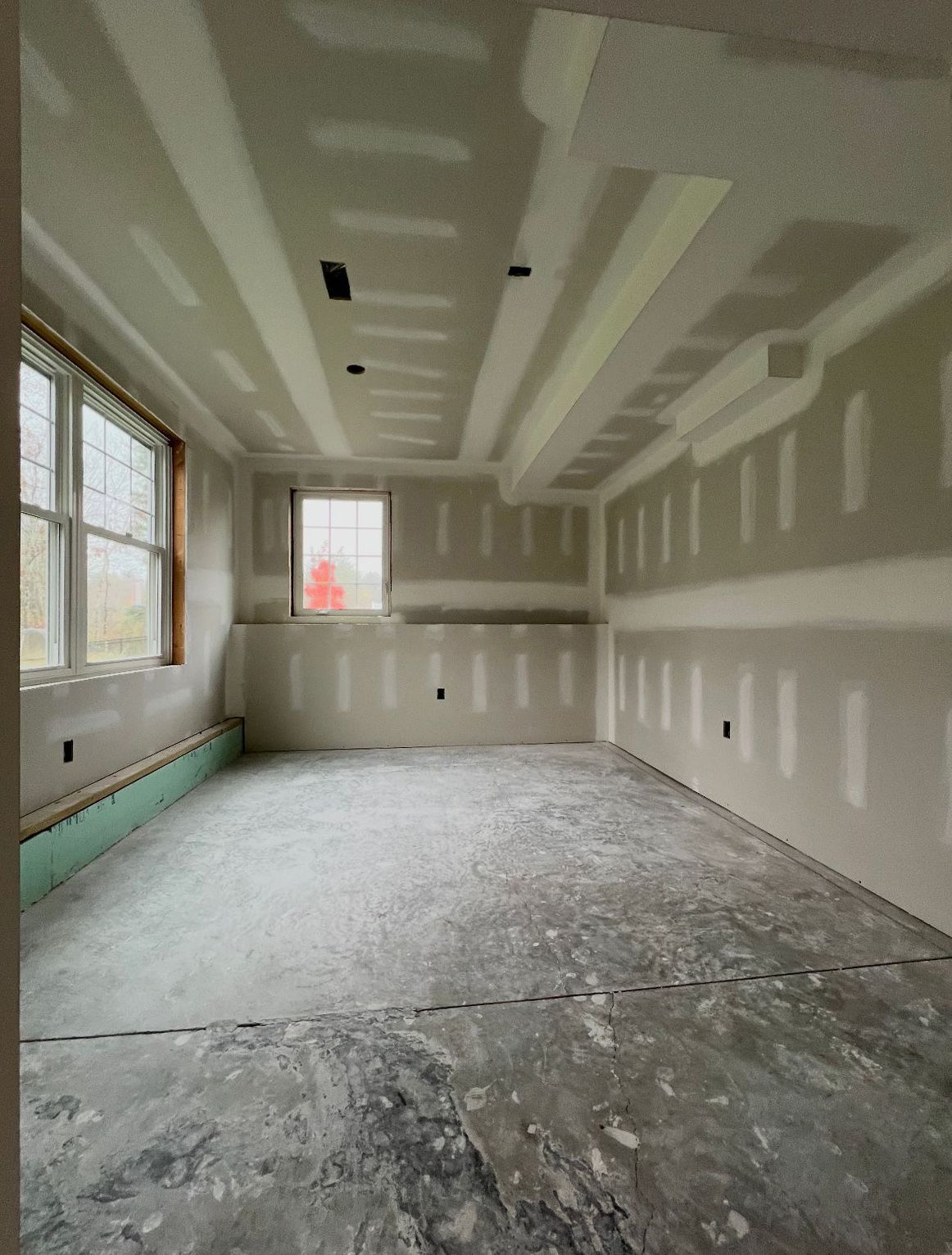 Interior of a room under construction, with drywall on walls and ceiling, unfinished concrete floor, and windows.