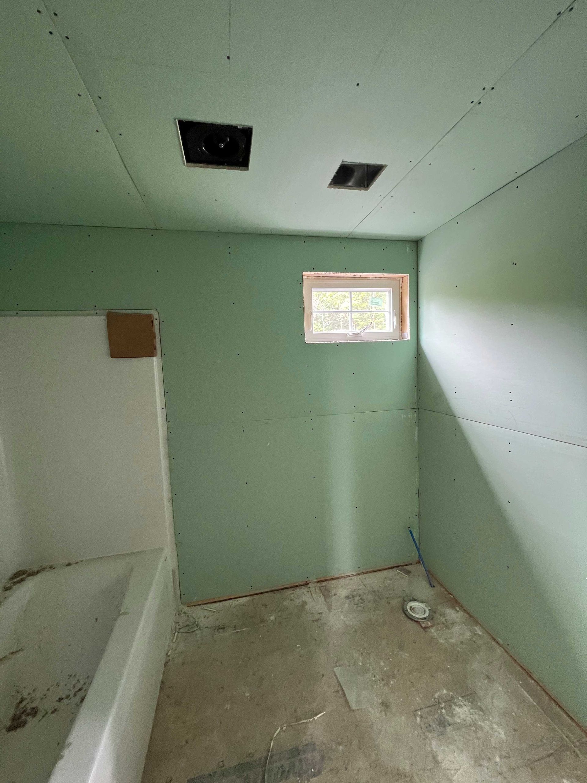 Unfinished bathroom with green drywall, a window, vent, and bathtub. The floor is concrete.