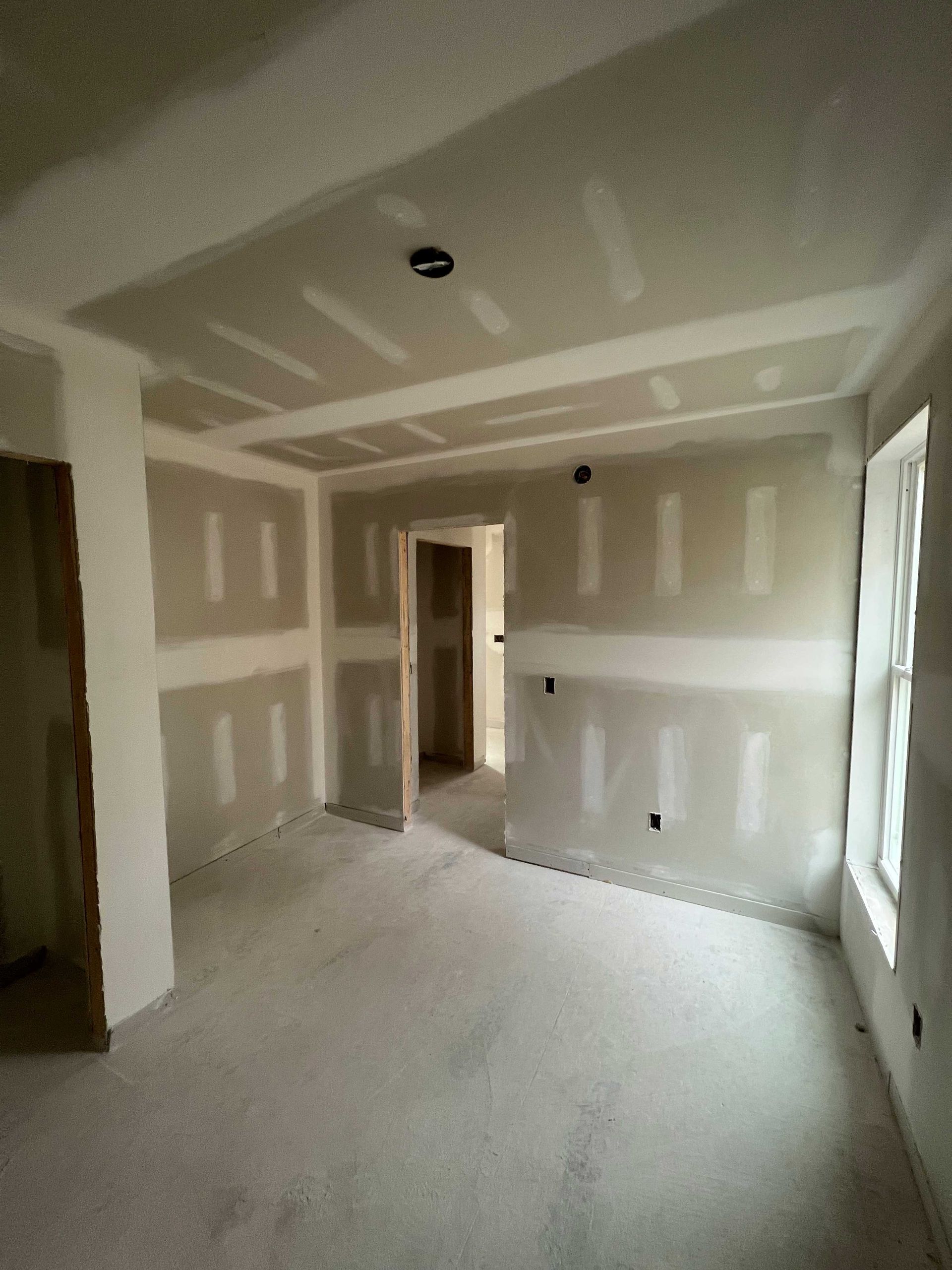 Interior of a room under construction, with drywall on walls and ceiling, unfinished floor, and an open doorway.