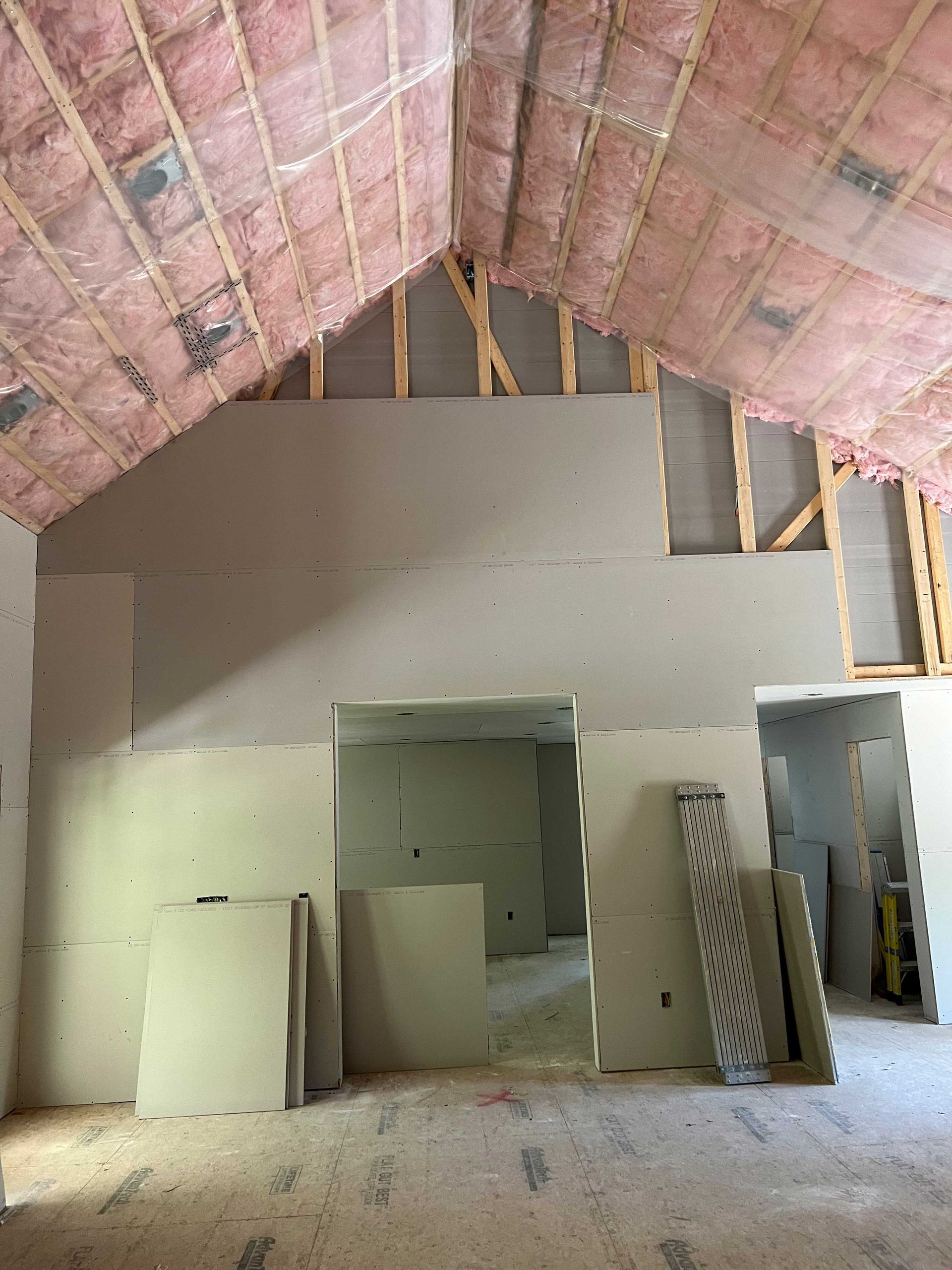 Interior view of a room under construction with exposed wood beams, pink insulation, and drywall.