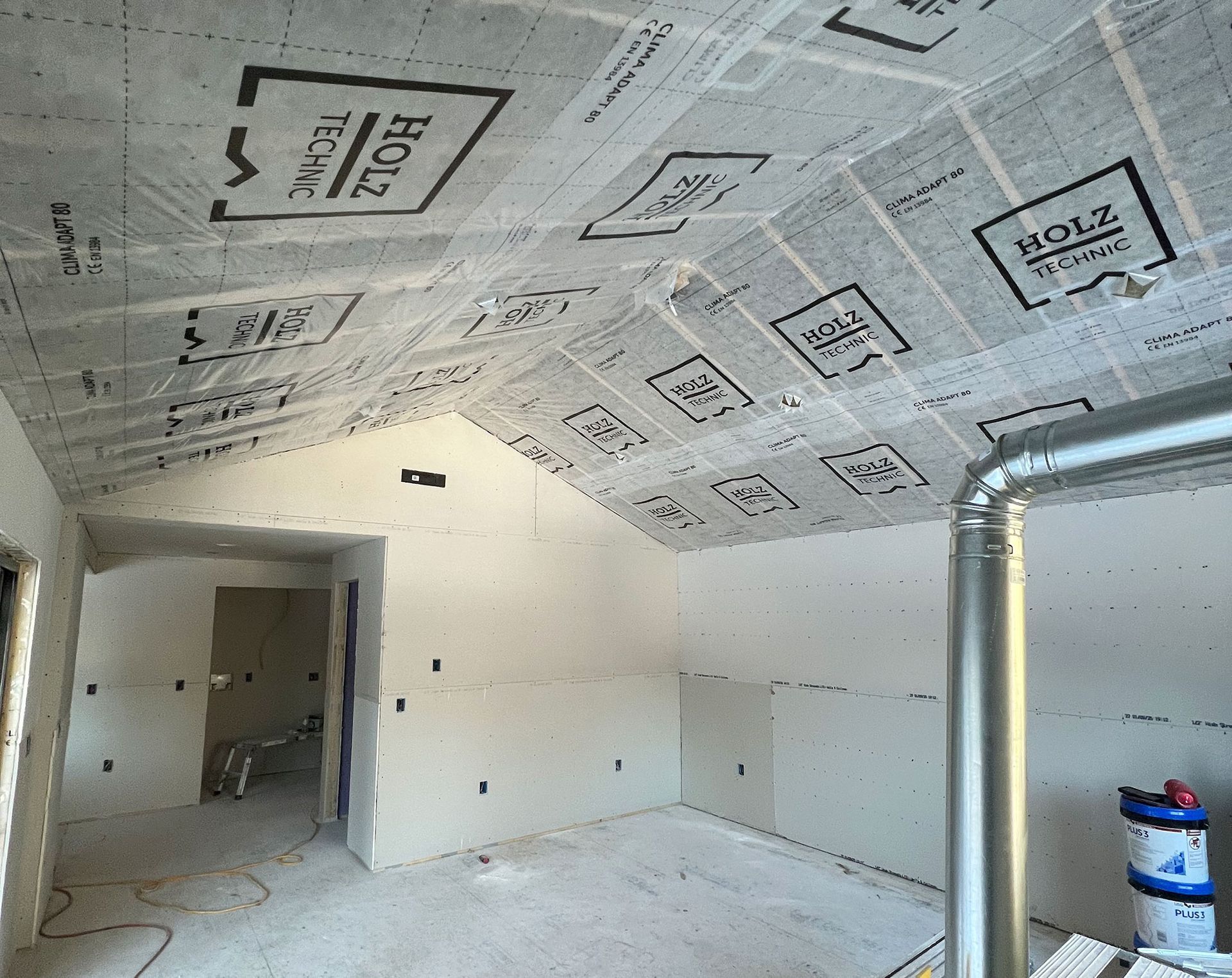 Interior of a room under construction, with drywall and ceiling membrane in place, and a metal vent pipe.