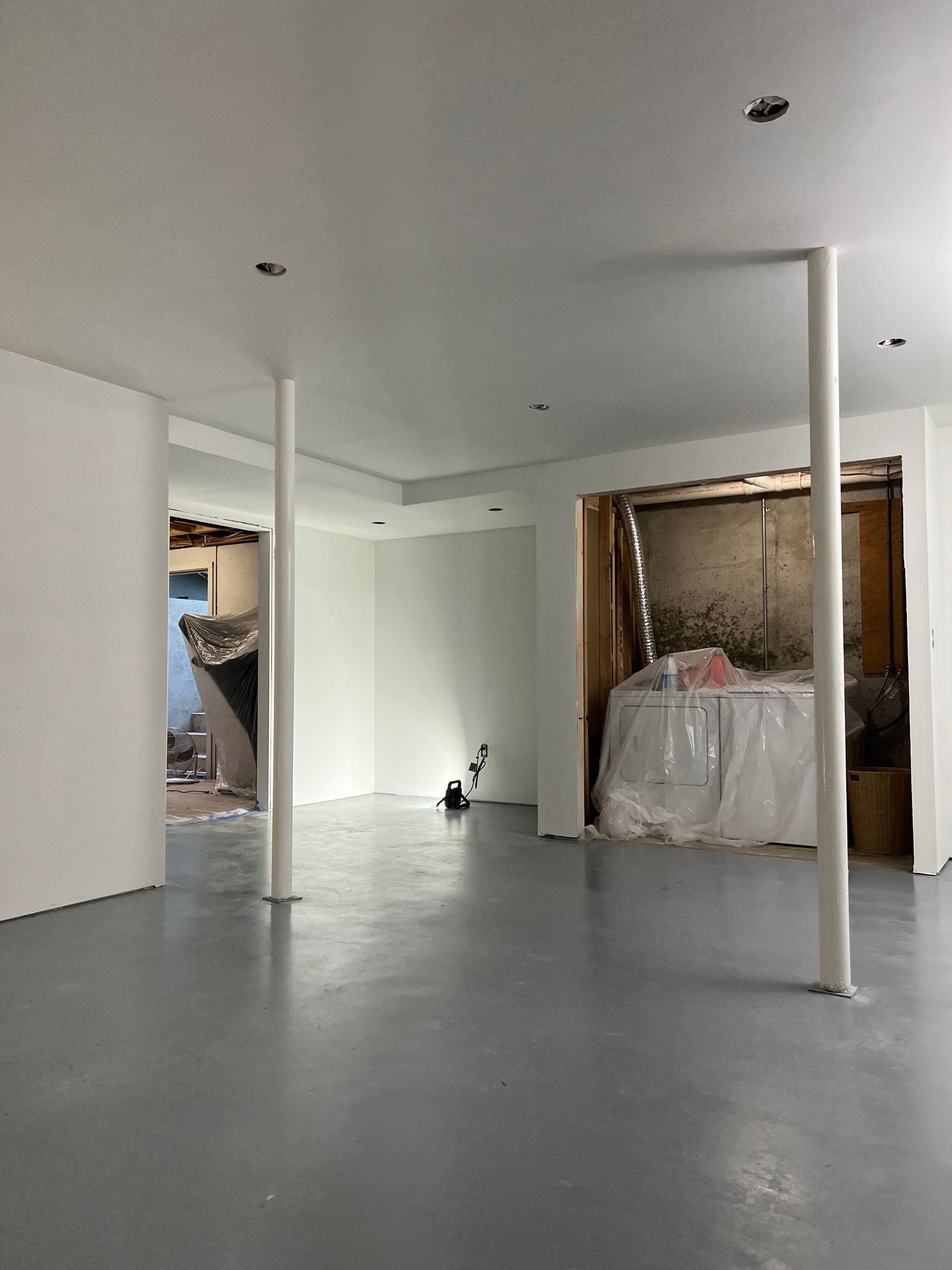 Empty unfinished room with white walls, gray floor, and exposed columns, with covered furniture at the back.