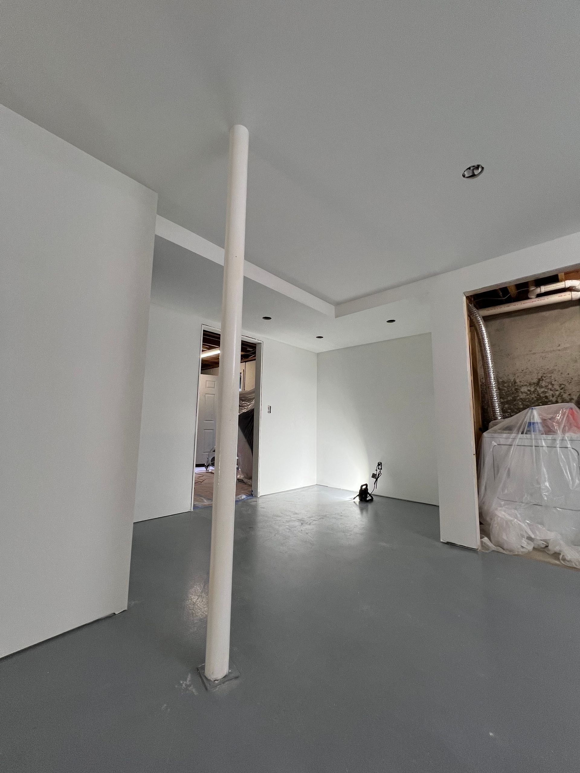 Empty white room with gray floor, a central support pole, and a window covered with plastic sheeting.