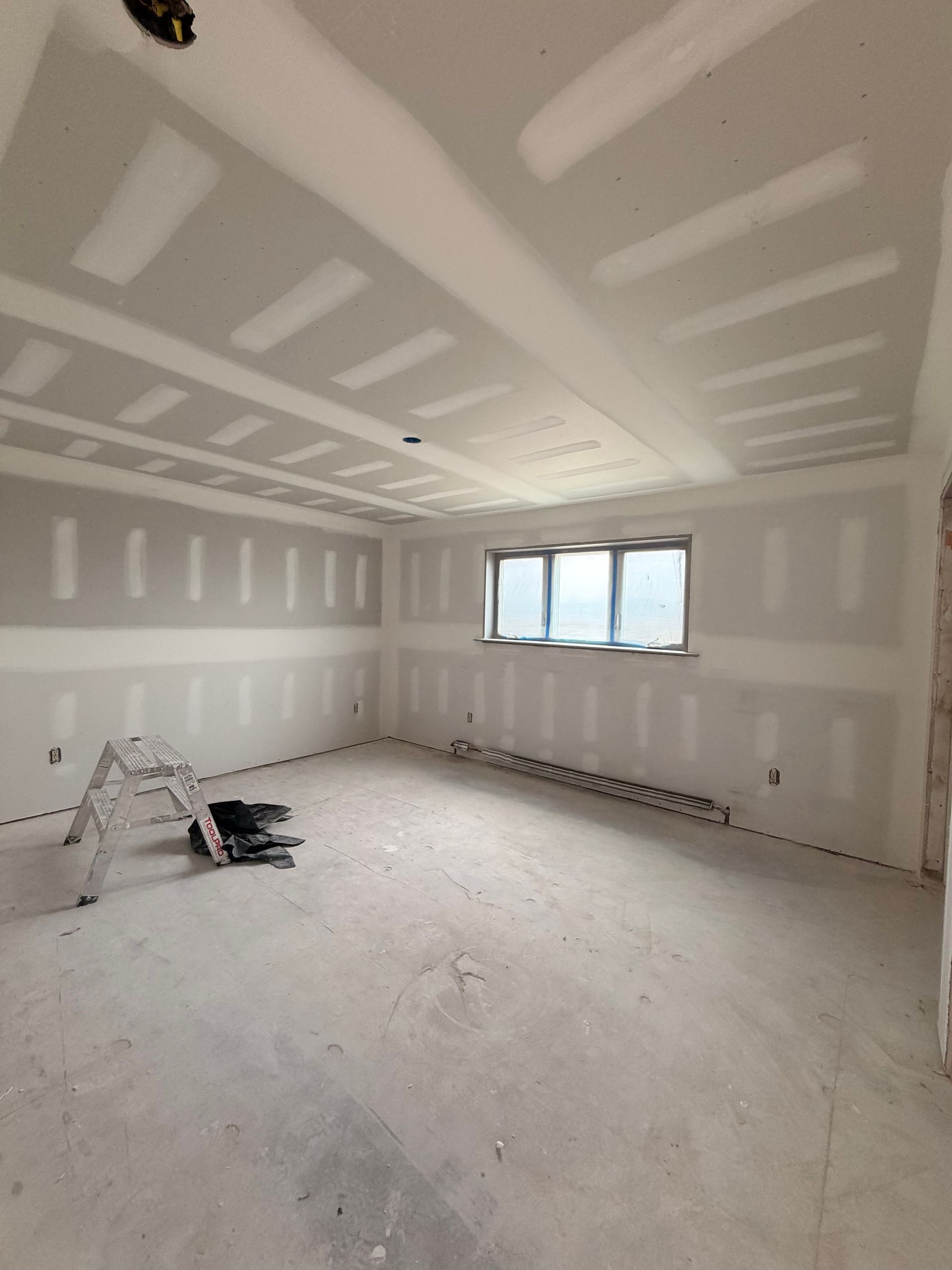 Unfinished drywall room with patchwork walls, ceiling, windows, and construction tools on the floor.