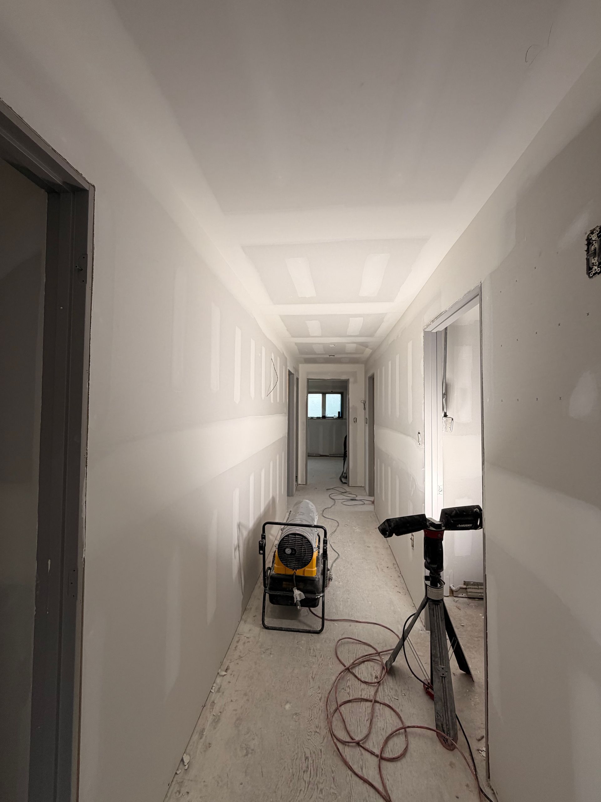 Narrow unfinished hallway with white walls, drywall dust, and construction tools on the floor.