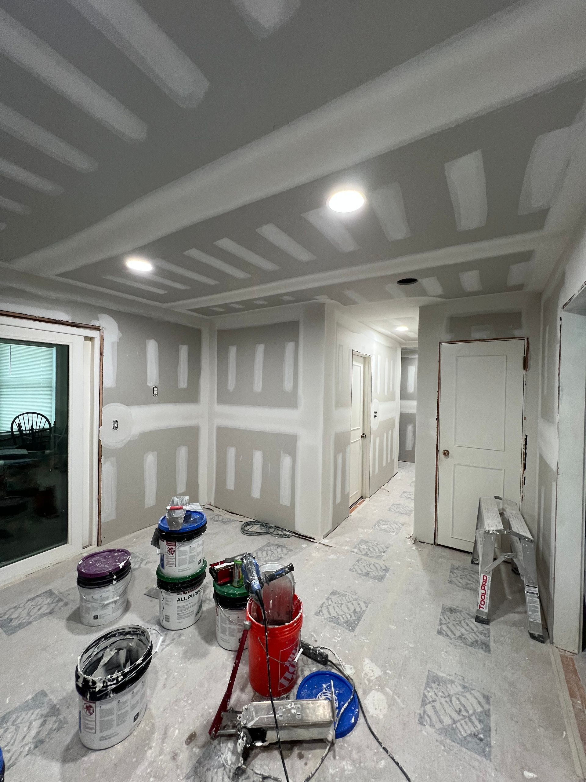 Drywalled room under renovation with paint buckets, tools, and a ladder on the floor.