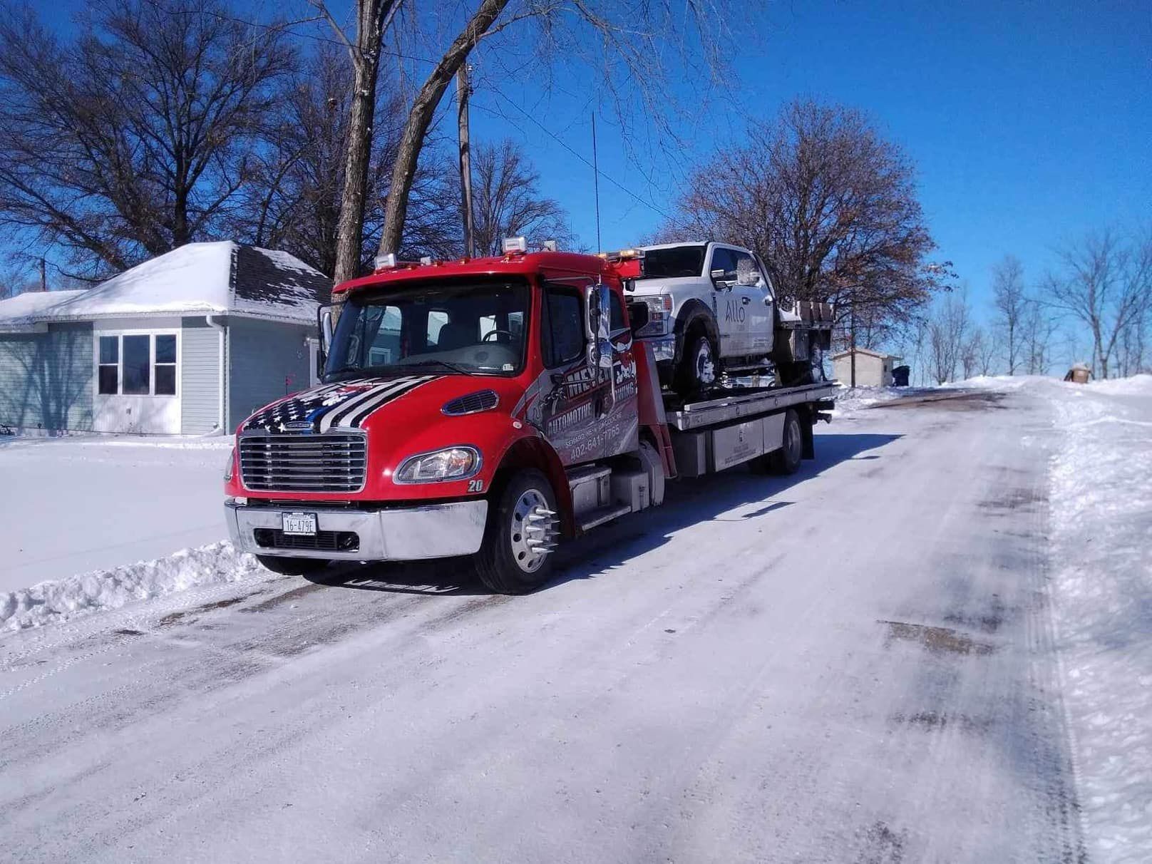 A red tow truck is carrying a white truck down a snowy road.