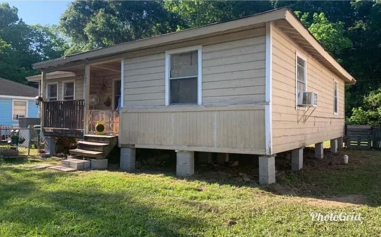 Small beige house on concrete blocks with a front porch and grassy yard on a sunny day