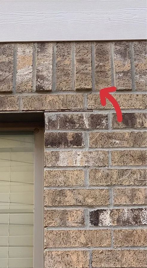 Brick house wall with a red arrow pointing to a vertical crack near the roofline.