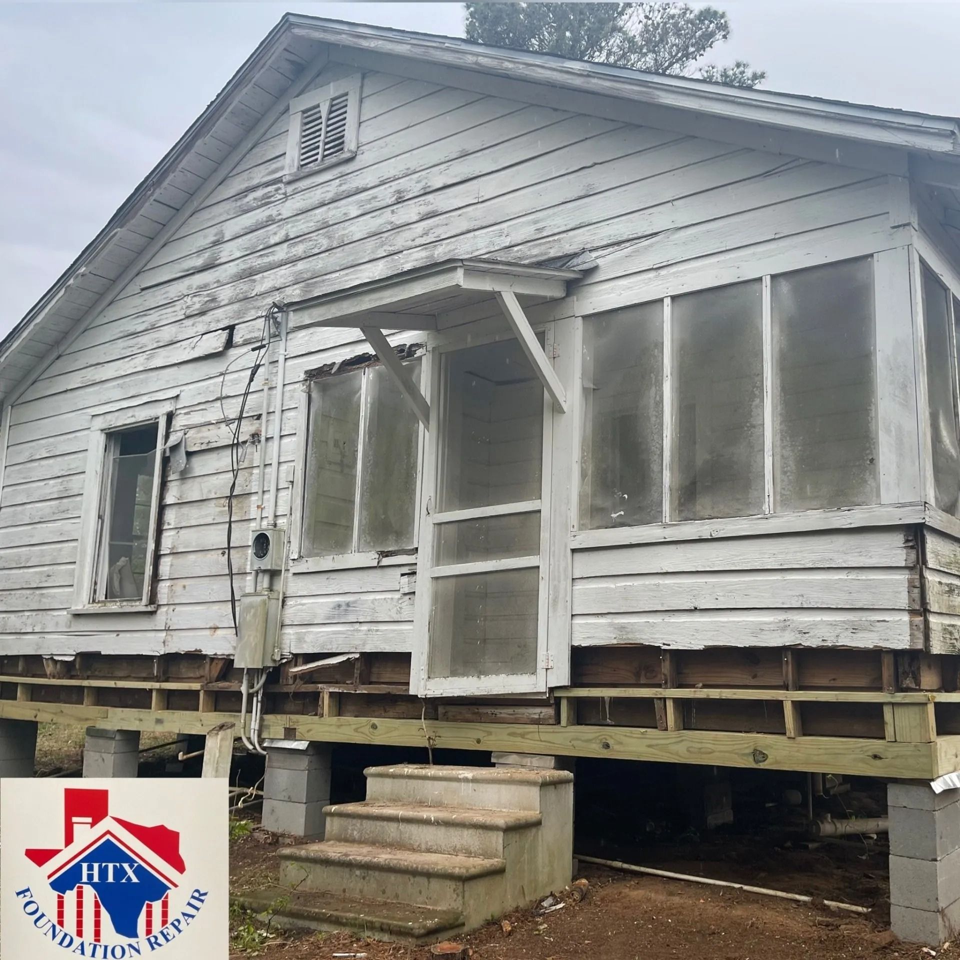 White house on raised supports with unfinished porch and boarded windows, showing exterior repair work.