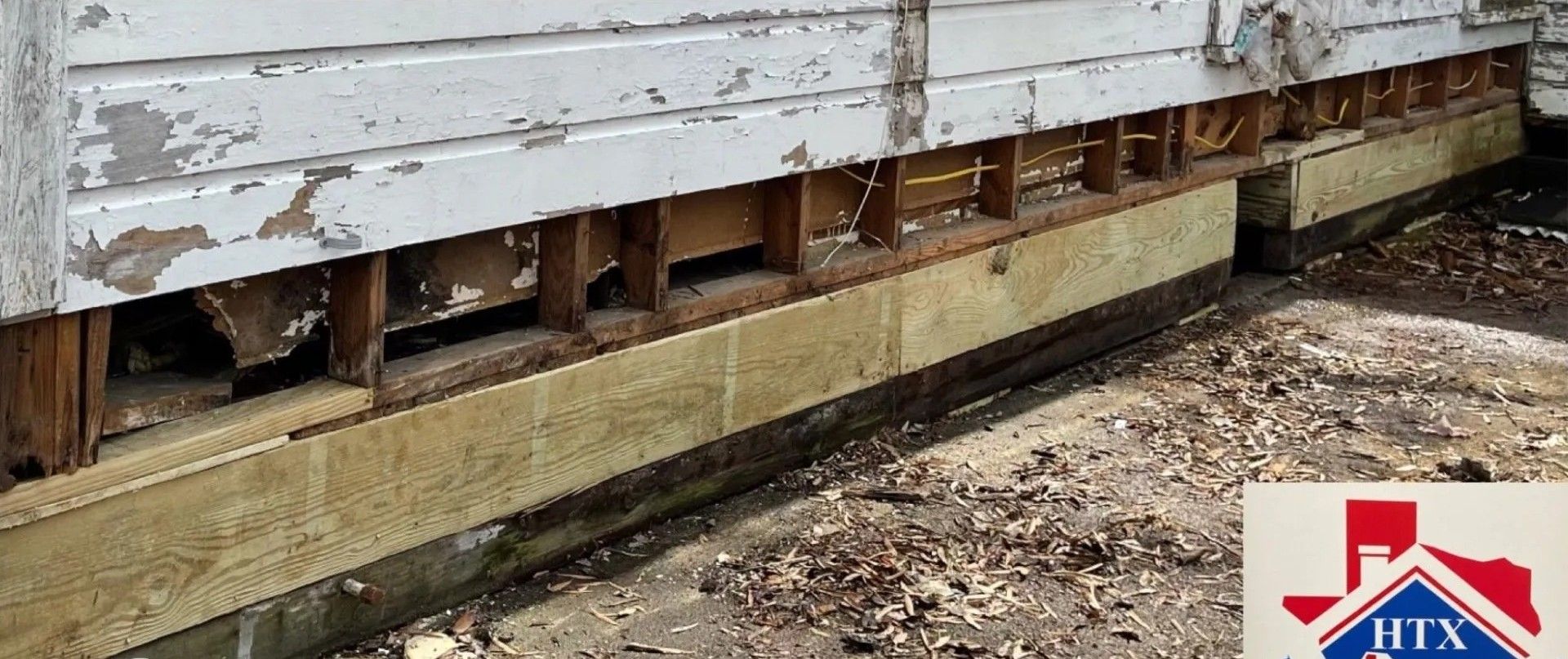 Peeling white house siding with exposed foundation and dirt ground below, showing exterior damage.