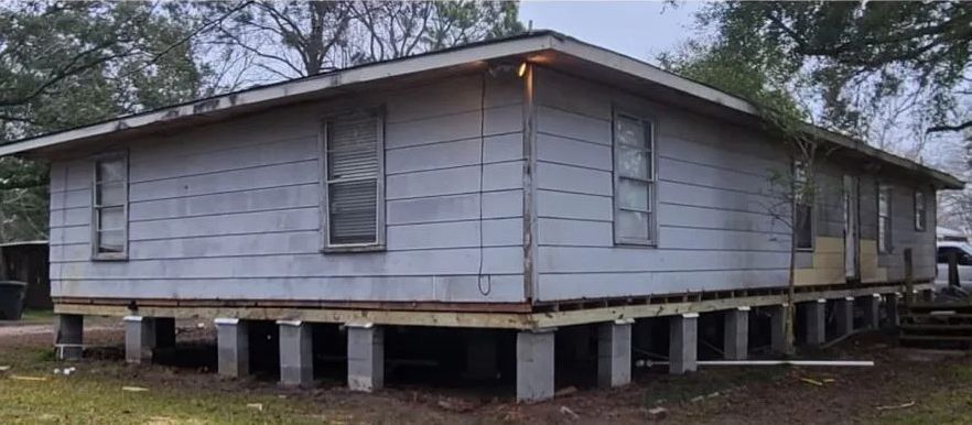 Elevated gray house on concrete piers with boarded windows and a covered porch in a wooded yard