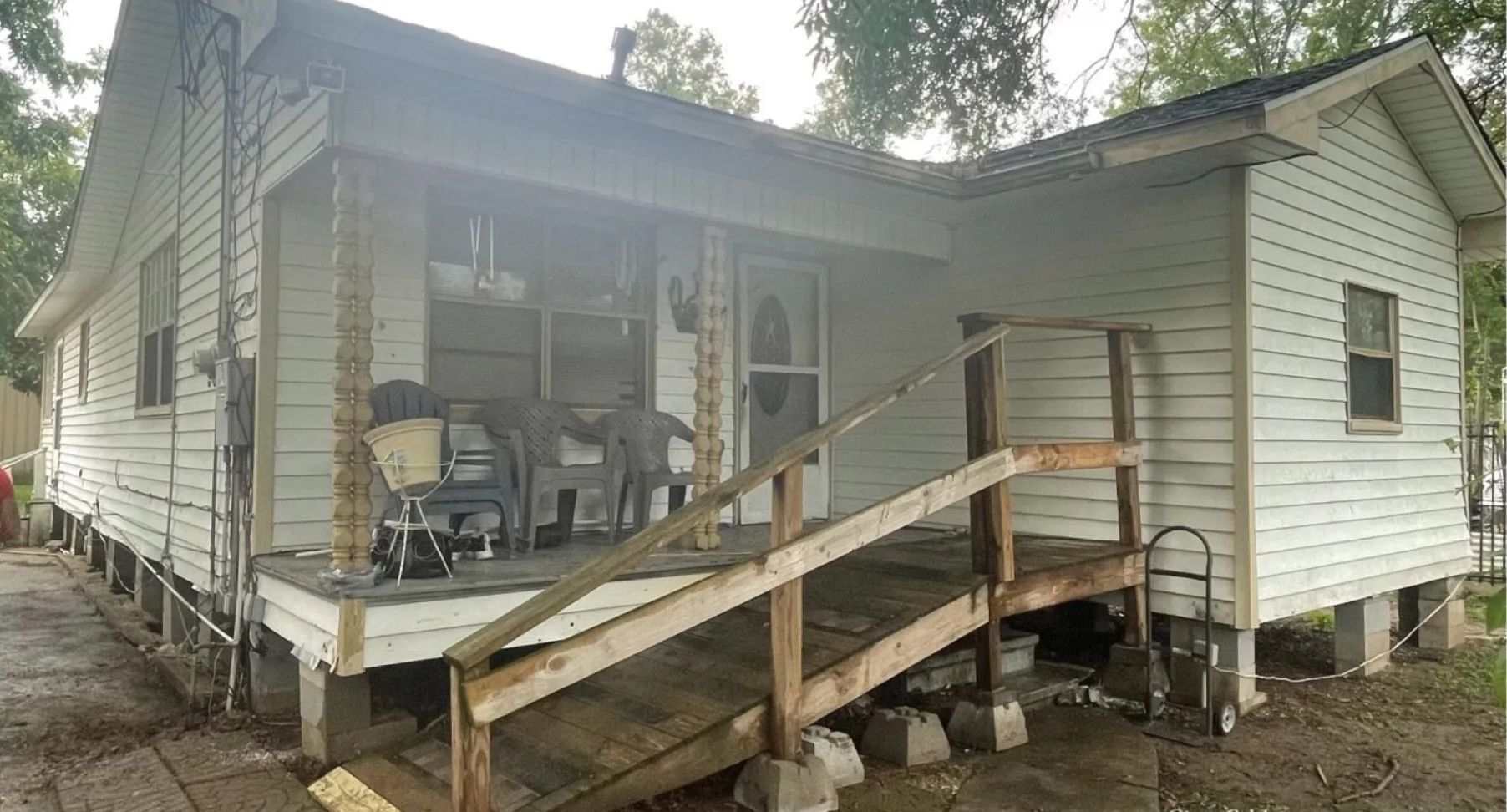 White house with a covered porch and wooden ramp, surrounded by trees and bare dirt yard