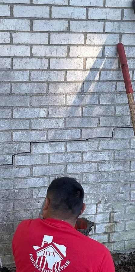 Worker in red shirt facing a gray brick wall with a long tool leaning at the right edge