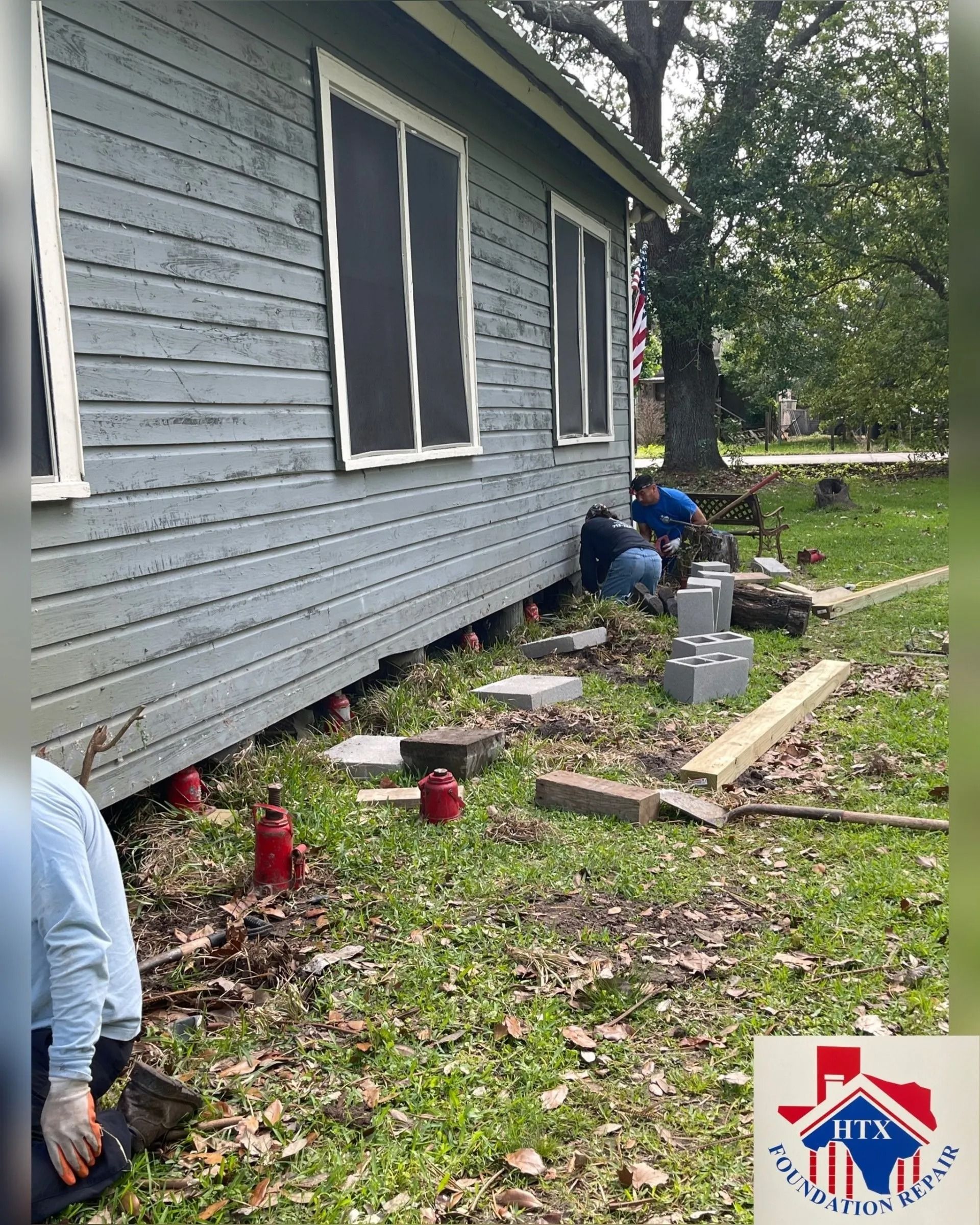 Gray house exterior with people working beside the foundation and scattered tools in the yard