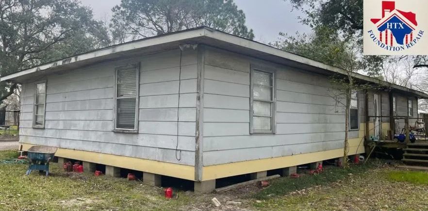 Gray single-story house on stilts with white trim and a red-roof logo in the top right corner