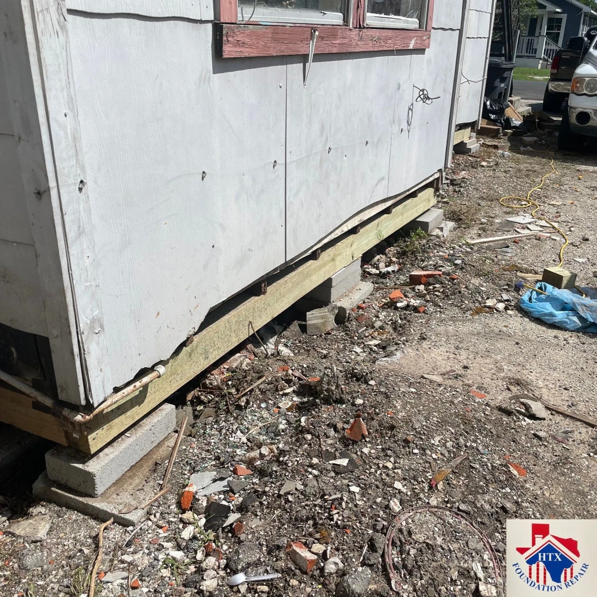 Side of a white building lifted on concrete blocks, with debris and gravel on the ground nearby.