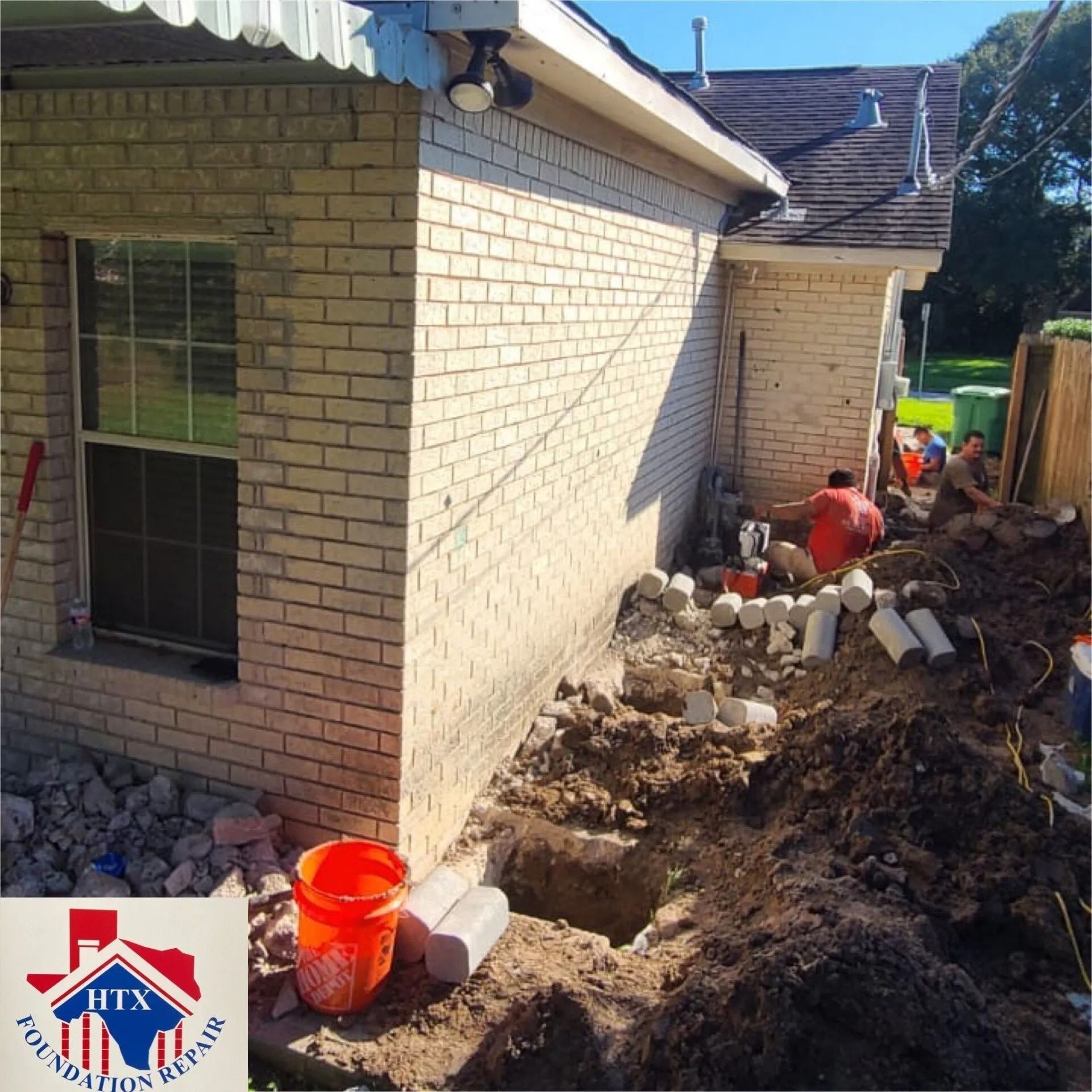 Backyard excavation beside a brick house, with workers digging near the foundation and rubble piled along the wall.