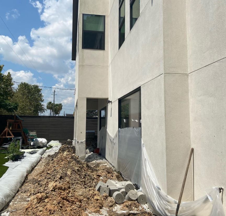 Construction site beside a white building with dirt, rubble, plastic sheeting, and snow along the path