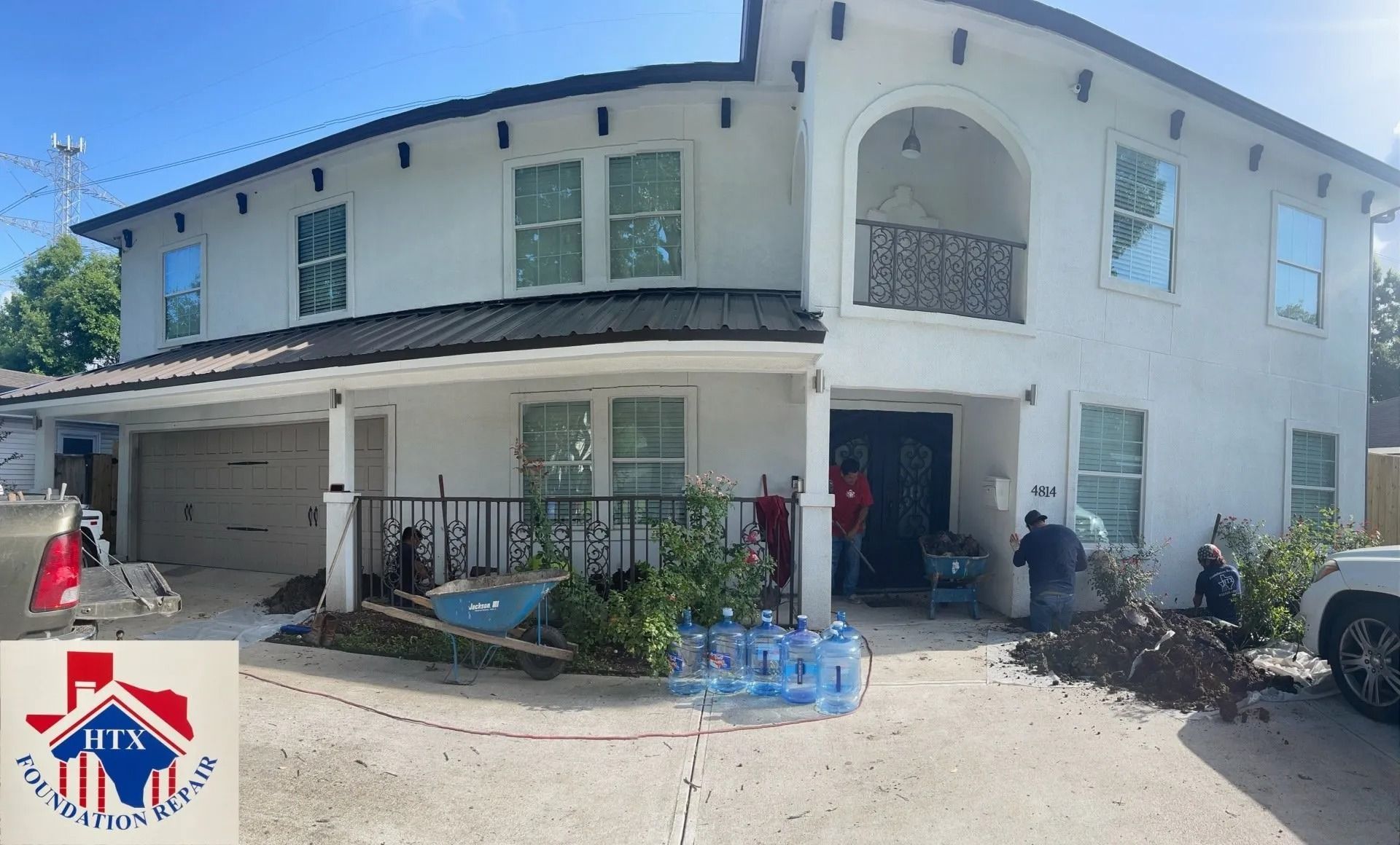 Two-story white house with balcony, parked cars, and people standing by the front entrance in bright daylight