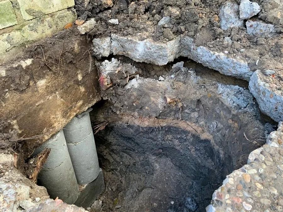 Excavated hole beside a stone wall with exposed pipes and dirt surrounding it