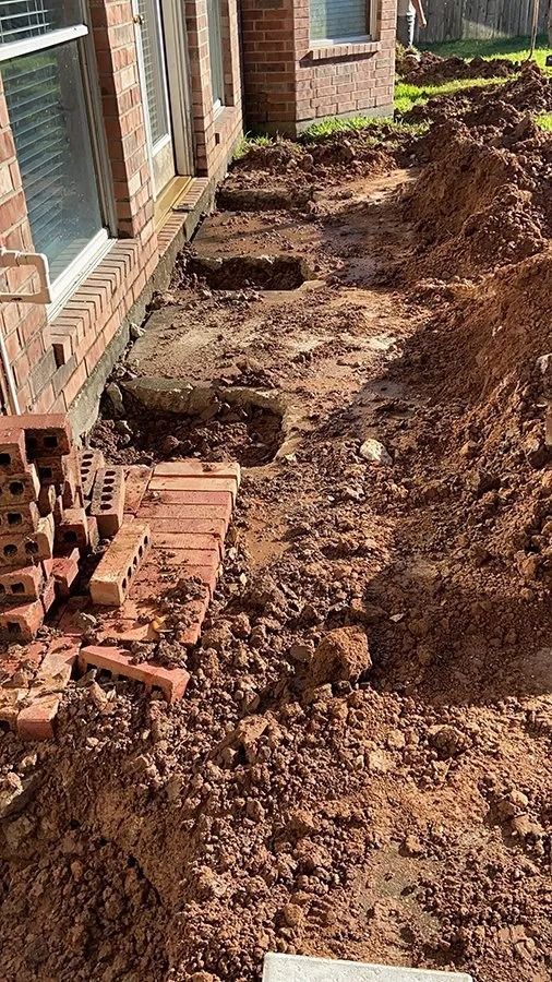 Freshly dug trench beside a brick house foundation, with piles of dirt and stacked bricks.