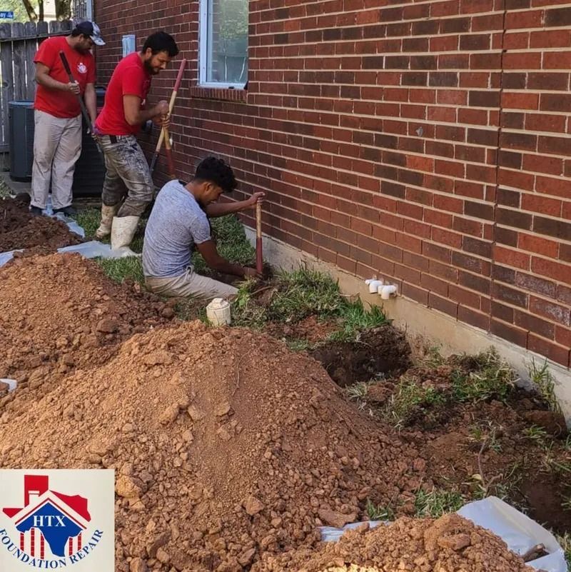 Workers digging trenches beside a brick wall, with piles of dirt and scattered plants along the foundation.