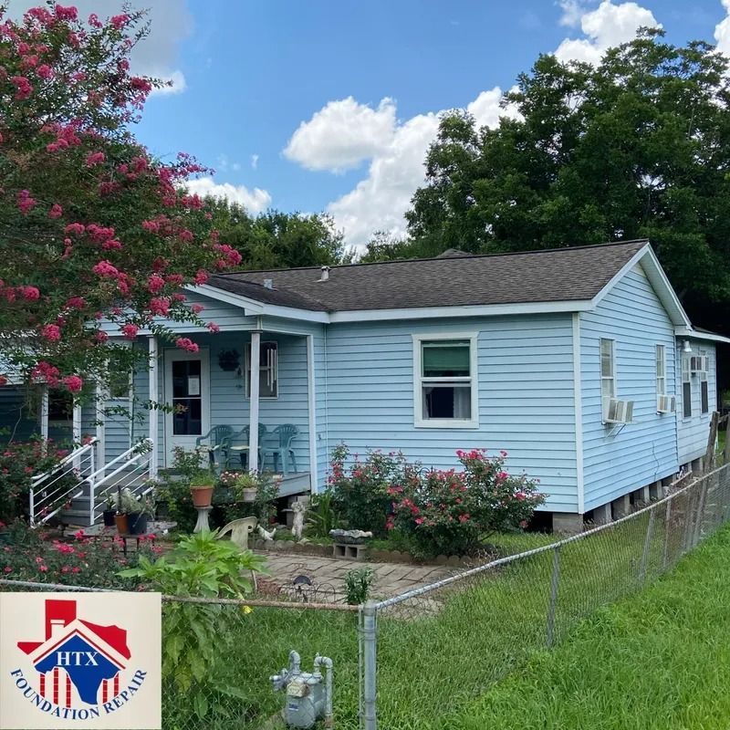 Light blue single-story house with white porch, pink flowers, and fenced yard under a sunny sky