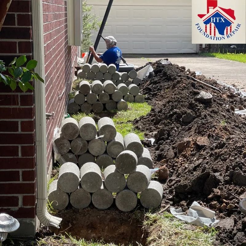 Worker stacking concrete blocks beside a brick house, with a dirt trench and soil pile nearby.