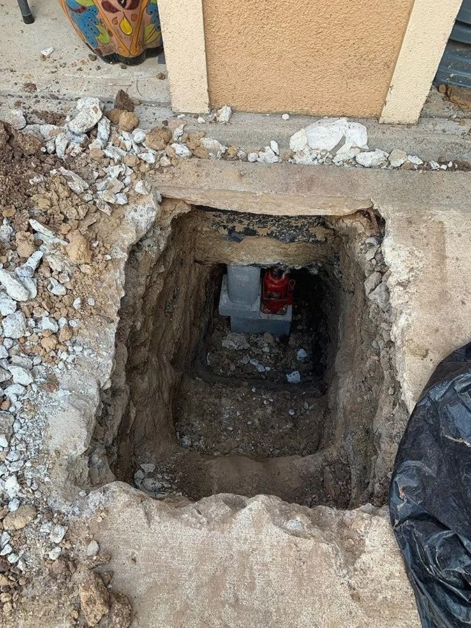 Excavated hole in concrete beside a building, with a red object visible inside.