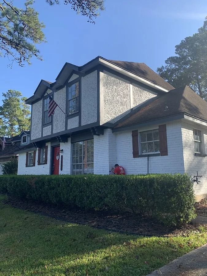 Two-story Tudor-style house with white siding, dark trim, red door, and trimmed hedges under a blue sky