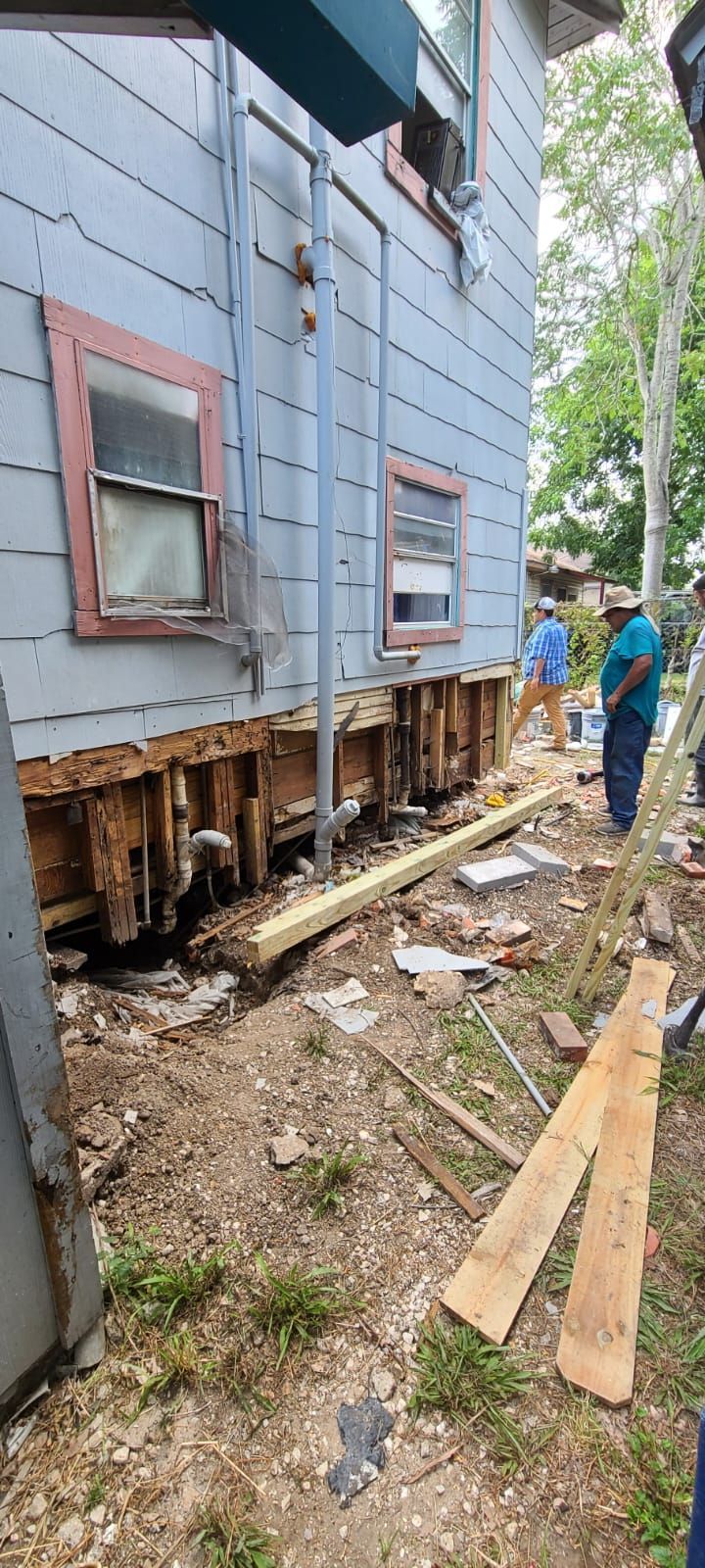 Workers inspect a house with a collapsed, exposed foundation and scattered debris after damage.