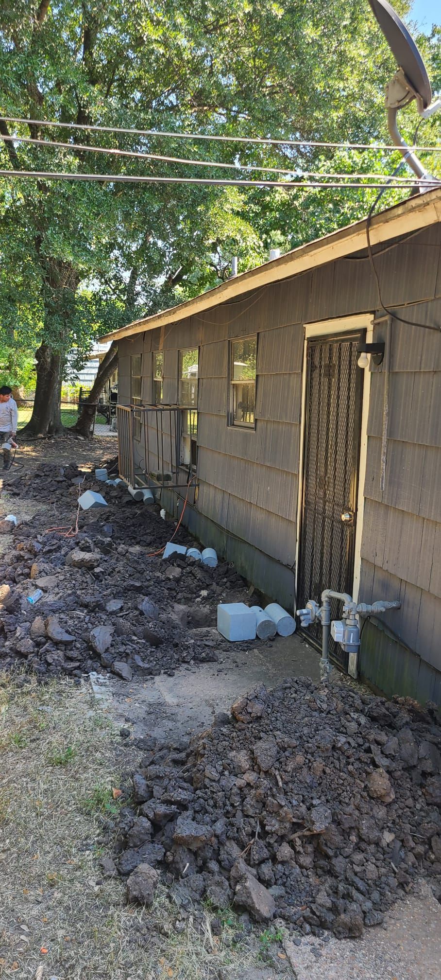 Mud-covered exterior of a damaged house with debris along the wall and trees overhead