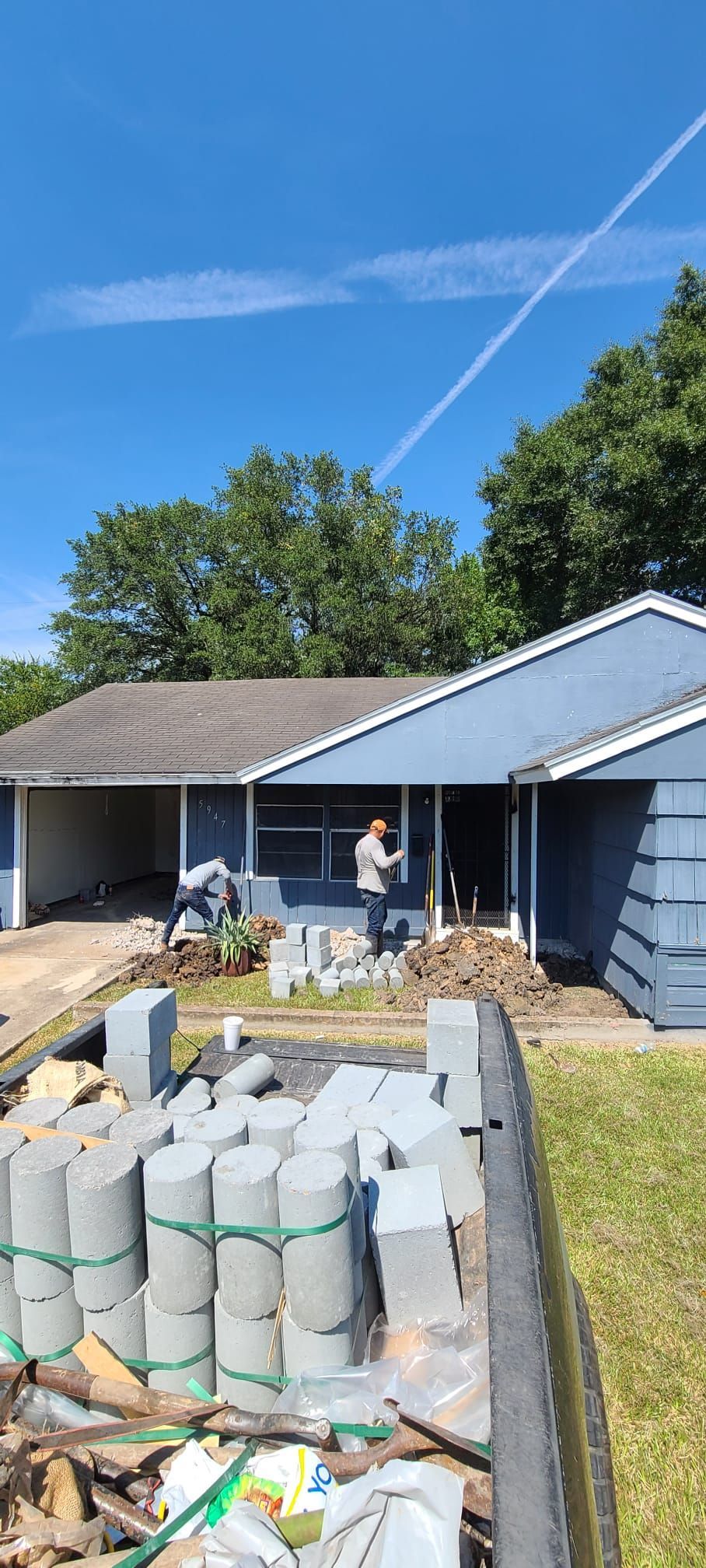 Person in a driveway beside stacked cinder blocks and a blue house under a clear sky