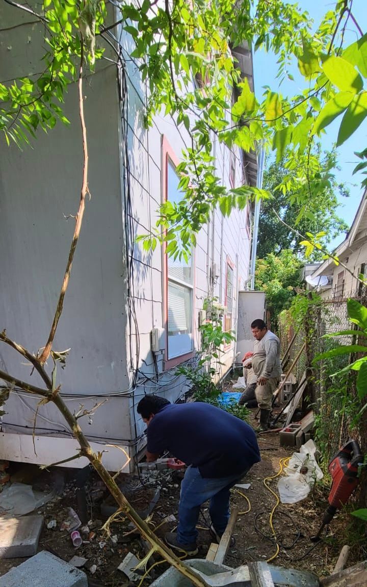 Two people clear debris beside a white building in a narrow, overgrown alley.